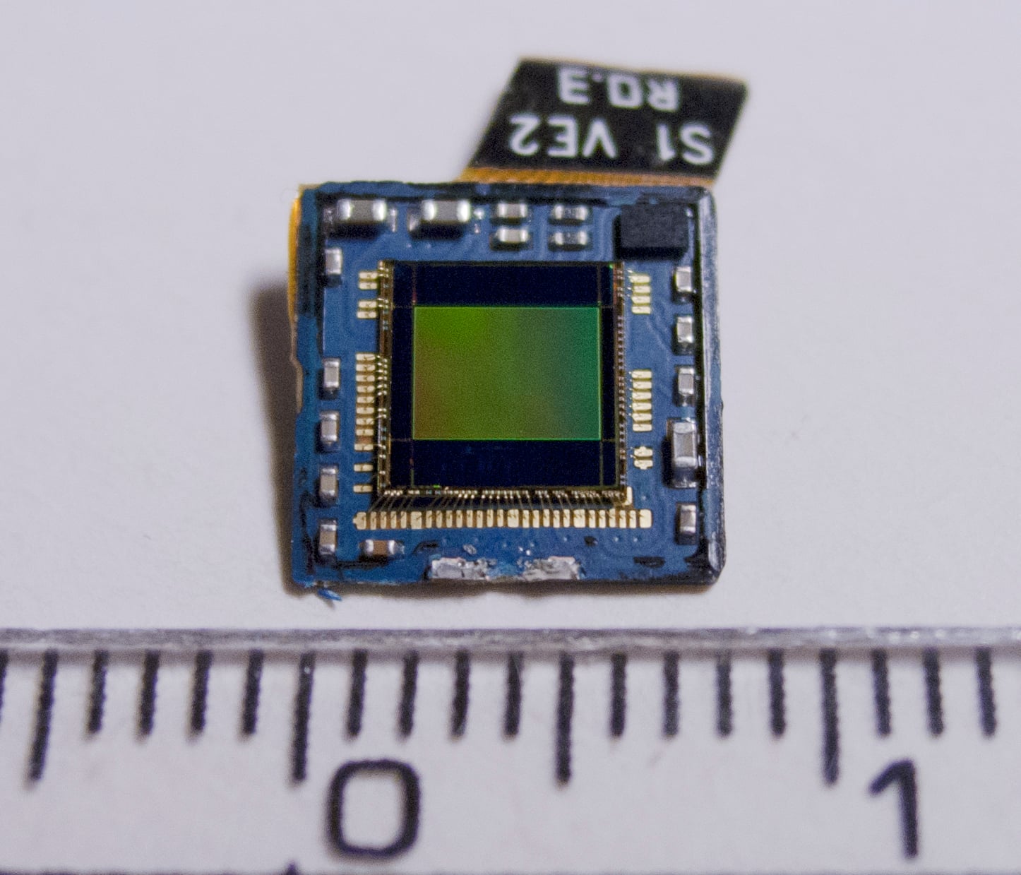 A small electronic sensor chip with a visible green and black square at the center, placed next to a ruler showing millimeter markings for scale.