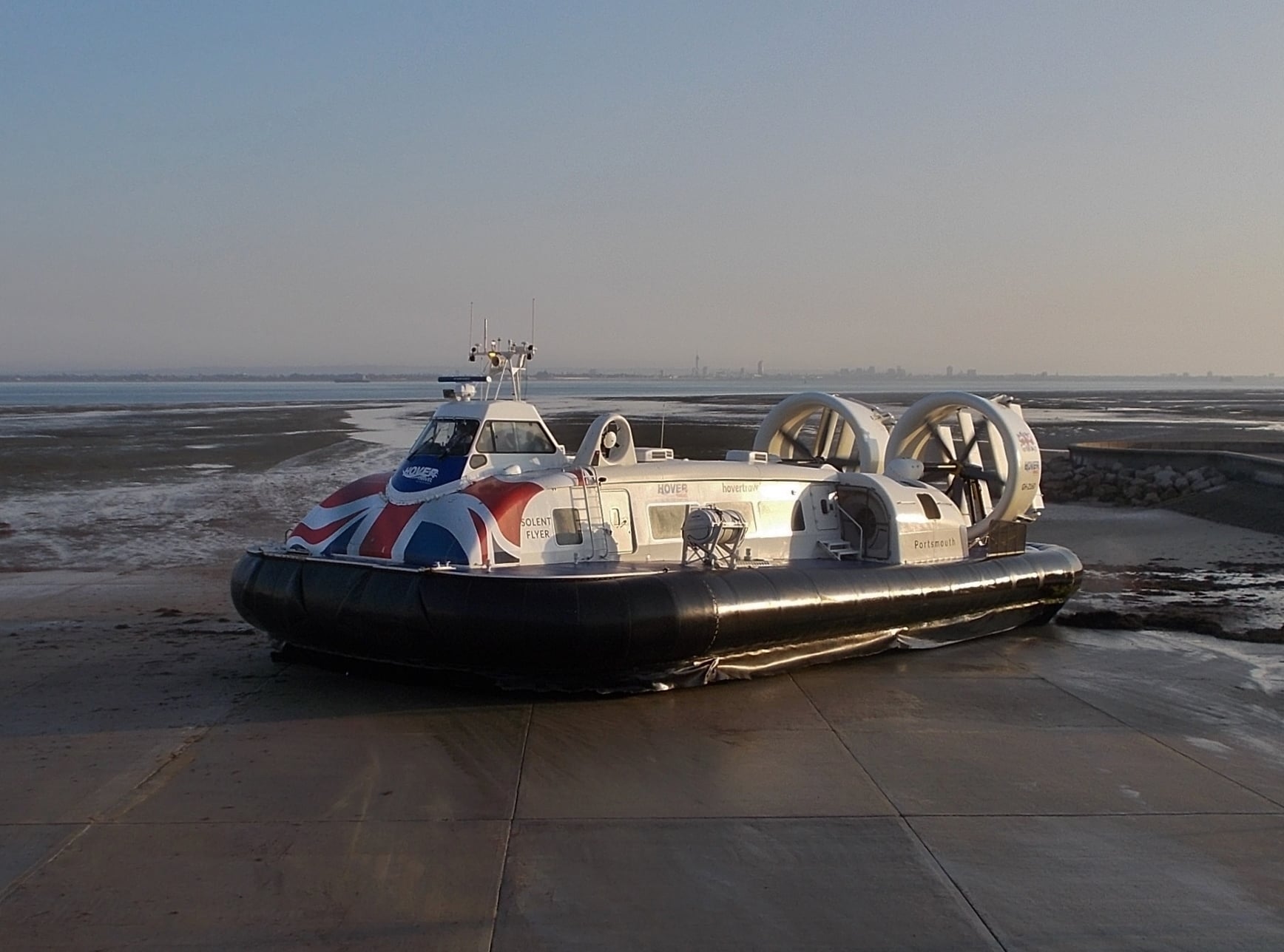 A white hovercraft with Union Jack flag graphics is parked on a concrete slipway near a sandy and muddy shoreline under a clear sky.