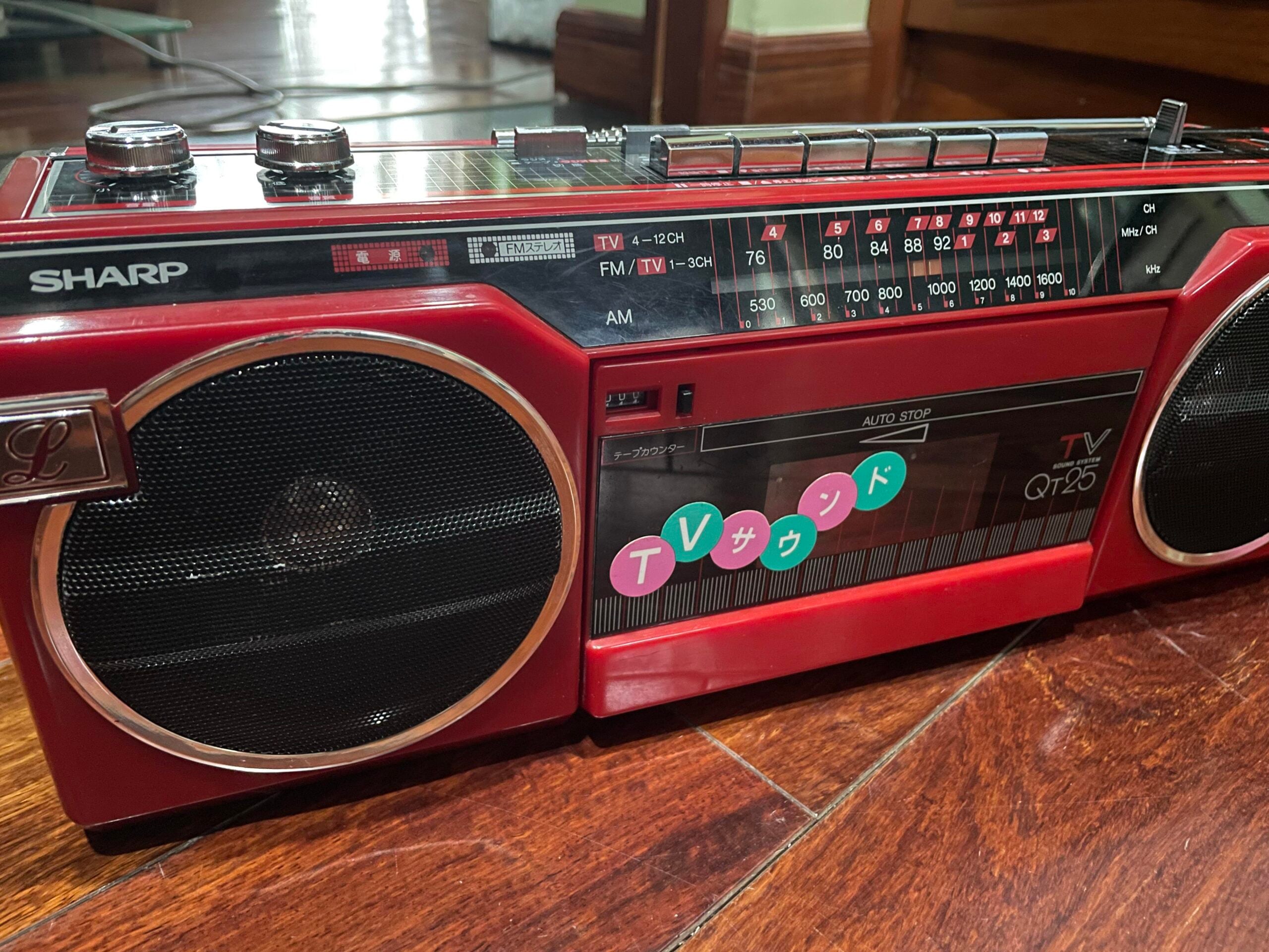 A red Sharp QT-25 vintage radio cassette player sits on a wooden floor, showing dials, speakers, frequency display, and colorful Japanese stickers on the front.