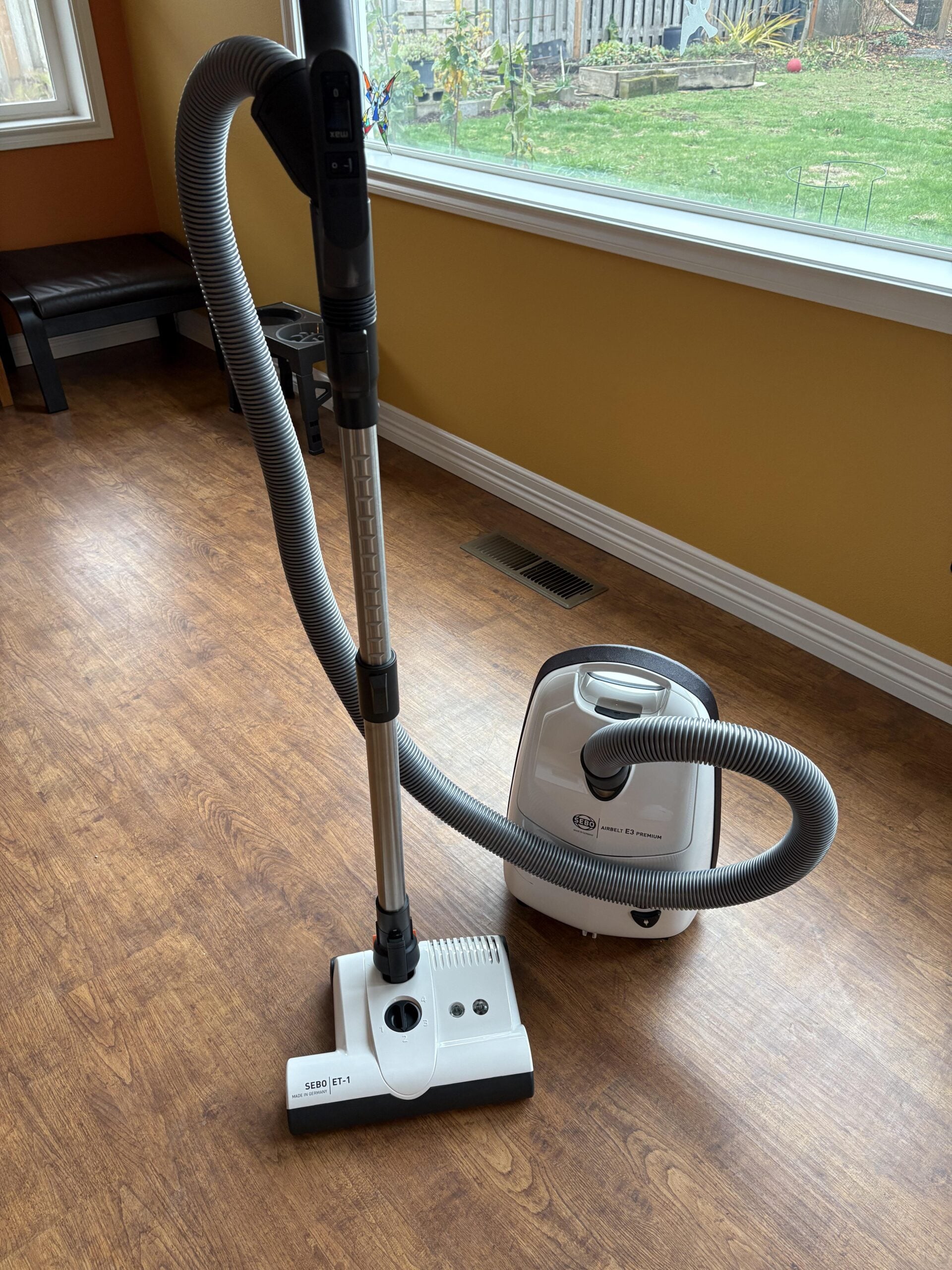 A canister vacuum cleaner with a hose and power head stands on a wood floor near a large window inside a room with yellow walls.