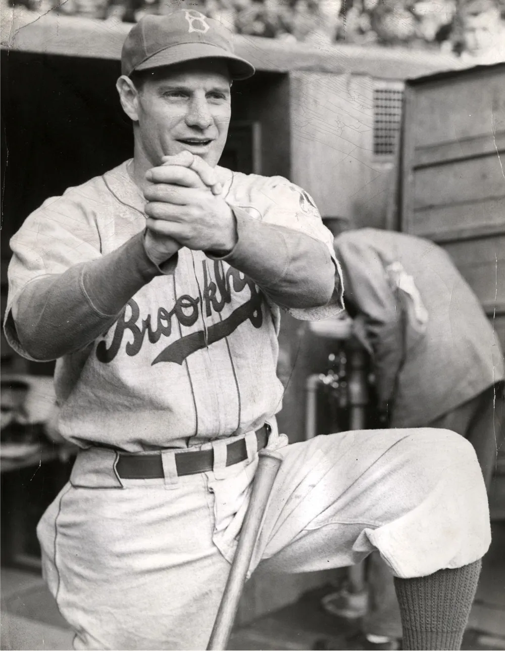 Baseball player in a “Brooklyn” uniform stands with one knee raised, hands clasped together, and a bat in front. Another person is in the background, bent over near an open shed or dugout. Black and white photo.