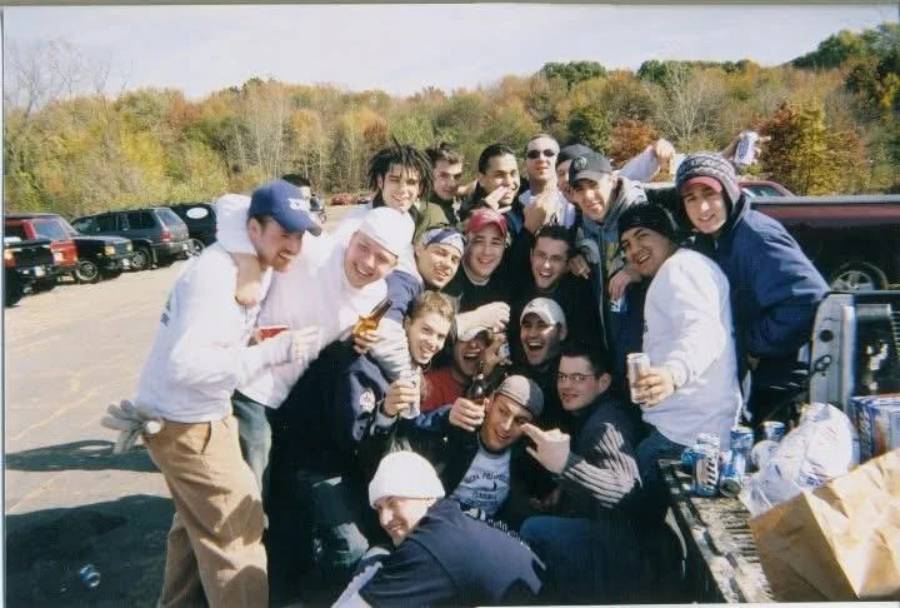A large group of young men poses together, smiling and laughing, on a sunny day near parked cars with trees in the background. Some hold drinks, and several are wearing hats and casual clothing.