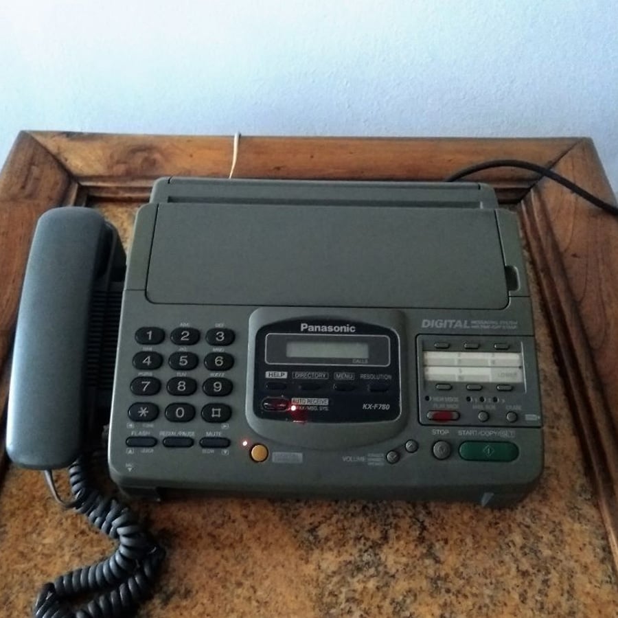 A Panasonic digital fax machine with a corded phone handset sits on a wooden table. The control panel displays buttons, lights, and a small screen.