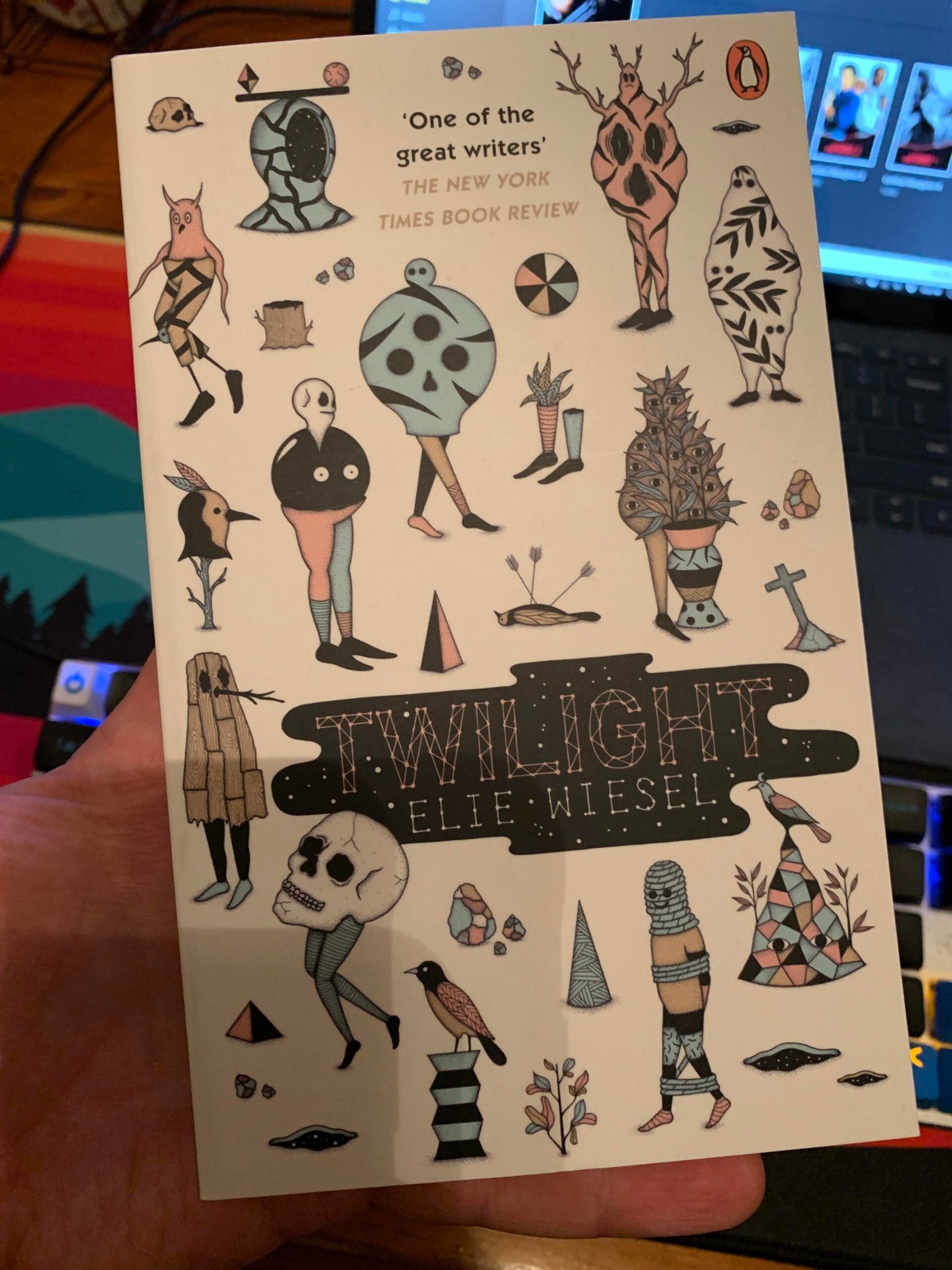 A hand holds the book “Twilight” by Elie Wiesel, featuring abstract, surreal illustrations of human and animal figures on the cover.