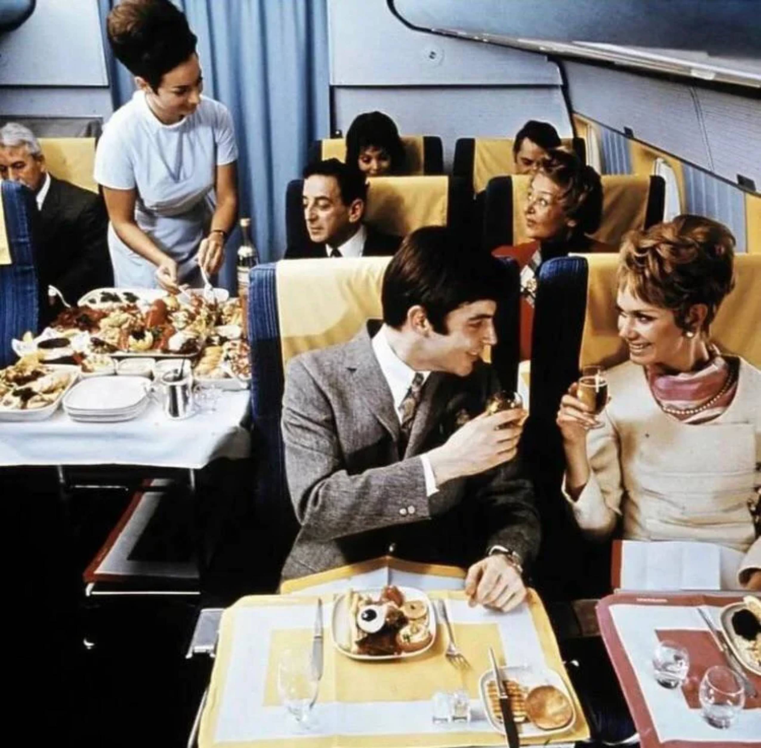 Passengers enjoy a meal and drinks in a vintage airplane cabin, with a flight attendant serving food. The seats have yellow headrests, and the atmosphere appears elegant and cheerful.