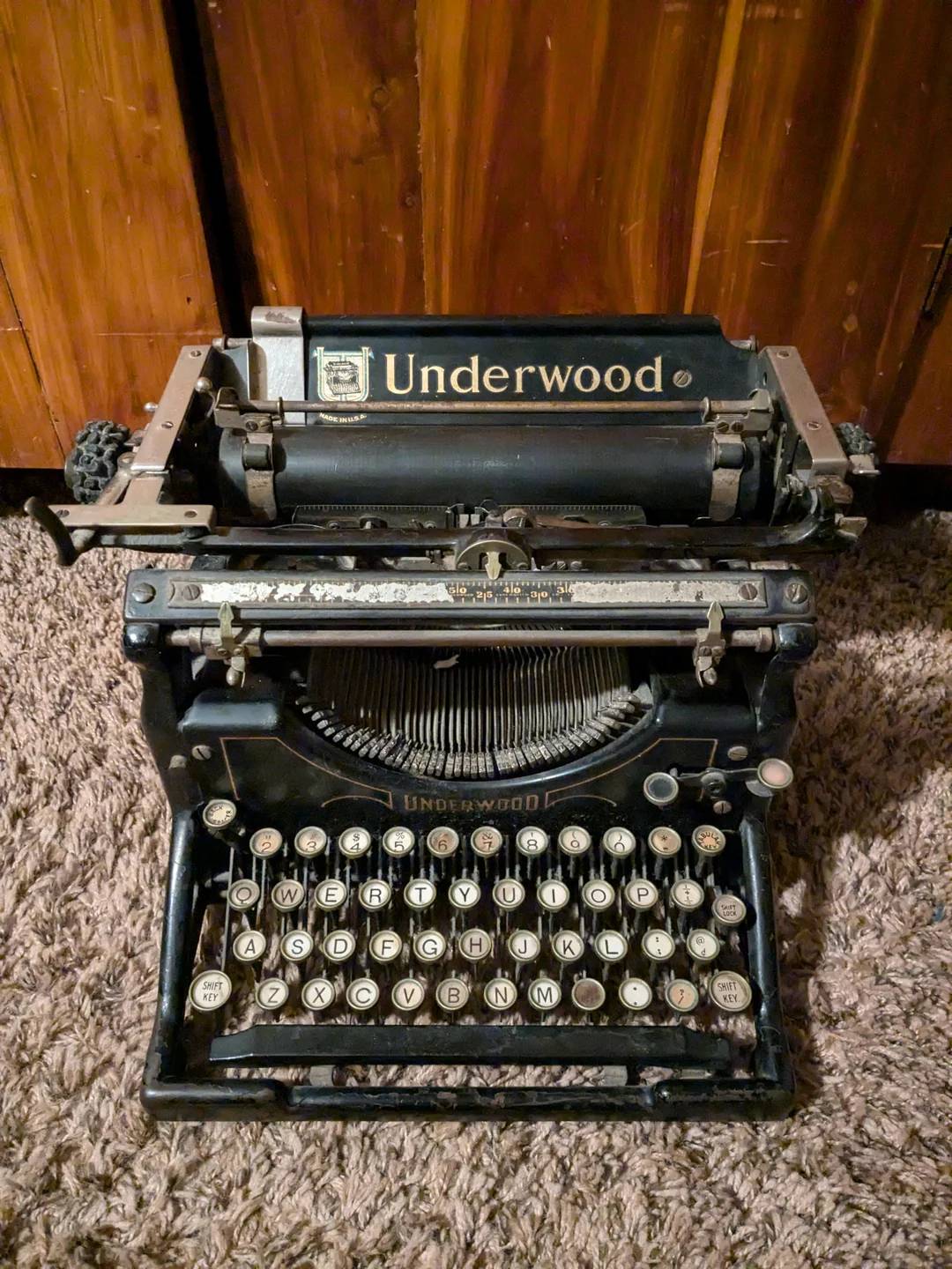 An antique Underwood typewriter sits on a carpeted floor in front of a wooden cabinet, with its keys and typebars exposed.