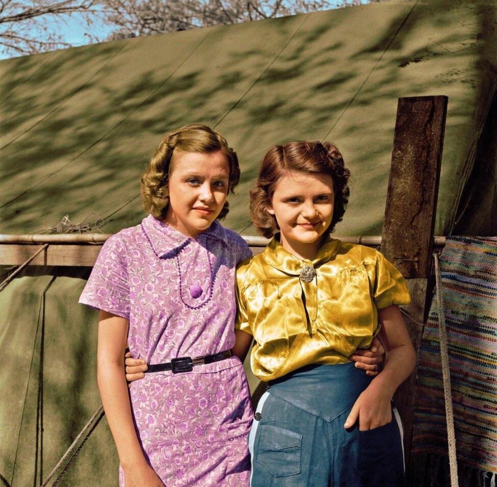 Two young women stand arm in arm, smiling in front of a green canvas tent. One wears a purple dress with a necklace, the other a shiny yellow blouse and blue skirt. The background includes trees and a woven mat.