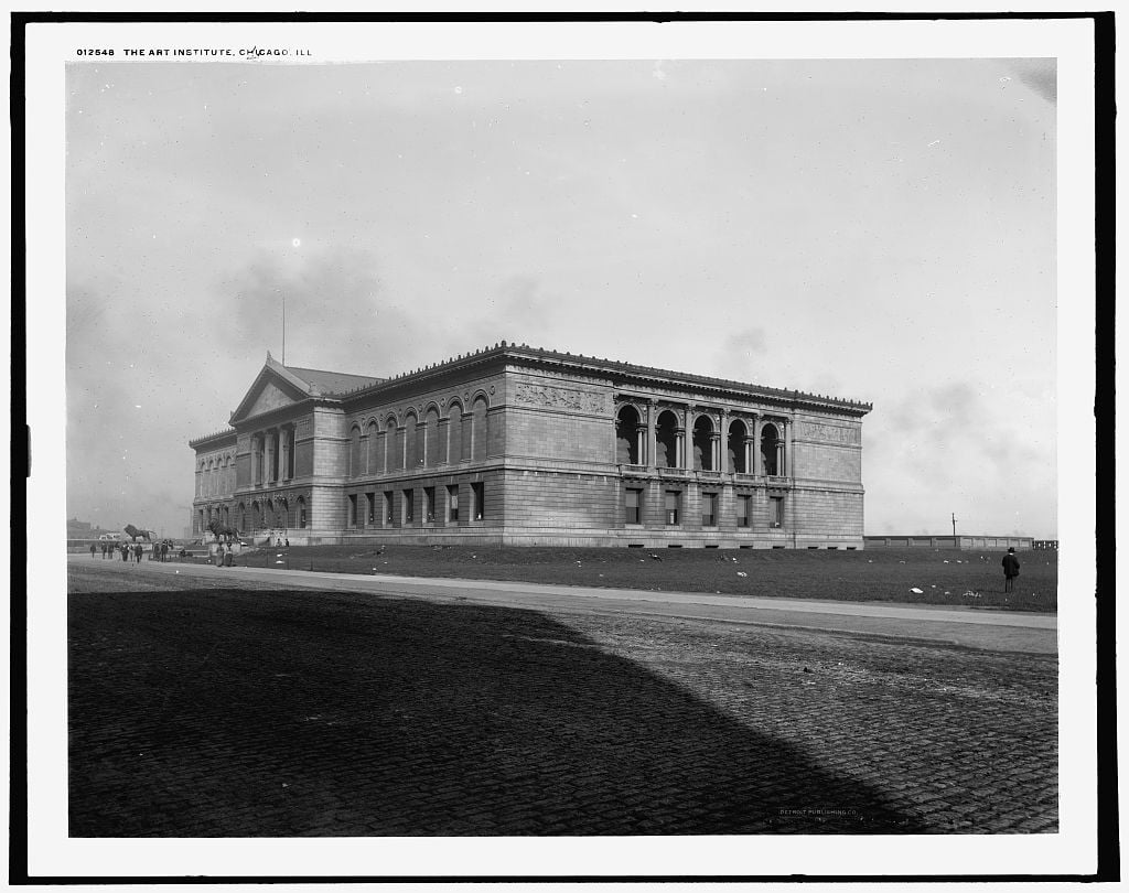 Historic black-and-white photo of the Art Institute of Chicago’s stone building. The street in front is cobblestone, with scattered pedestrians and an open, grassy area surrounding the structure.