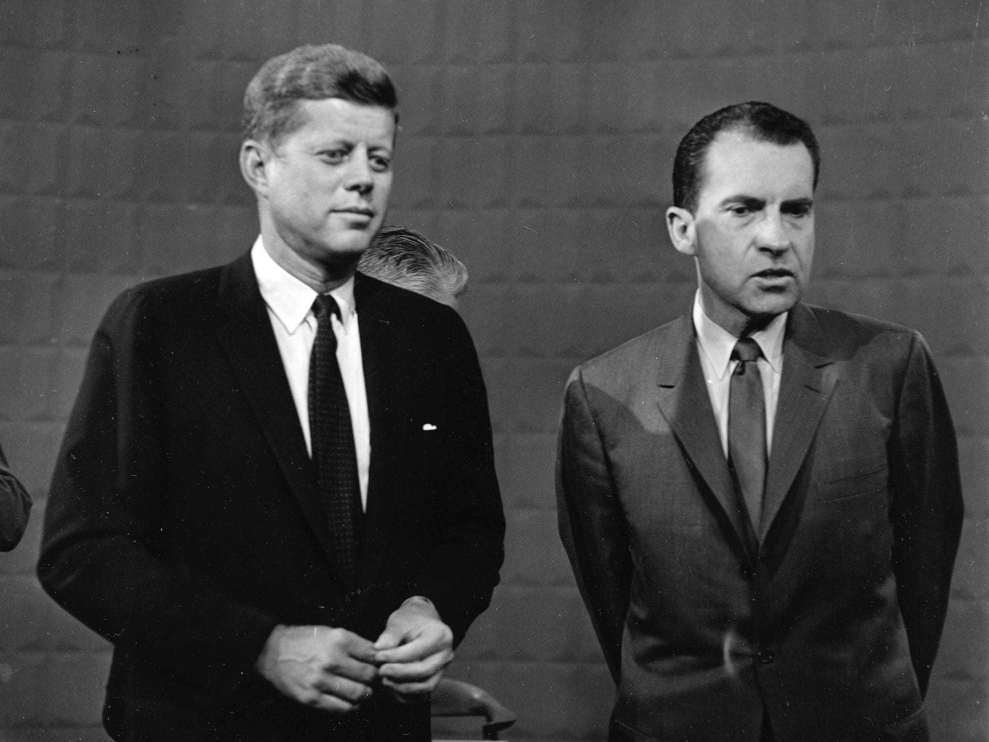 Two men in suits stand side by side in front of a plain, textured background, appearing serious and focused.