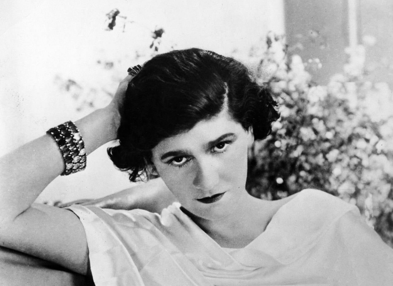 A woman with short dark hair in a white top leans her head on her hand, looking directly at the camera. She wears stacked bracelets and is posed in front of a blurred floral background.