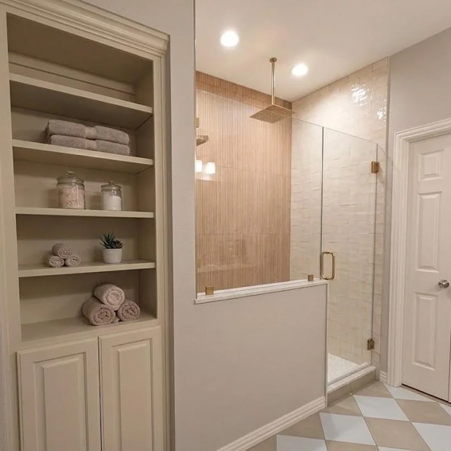 Modern bathroom with built-in shelves holding folded towels, glass jars, and a small plant beside a glass shower enclosure with a rain showerhead. Neutral beige tones throughout.