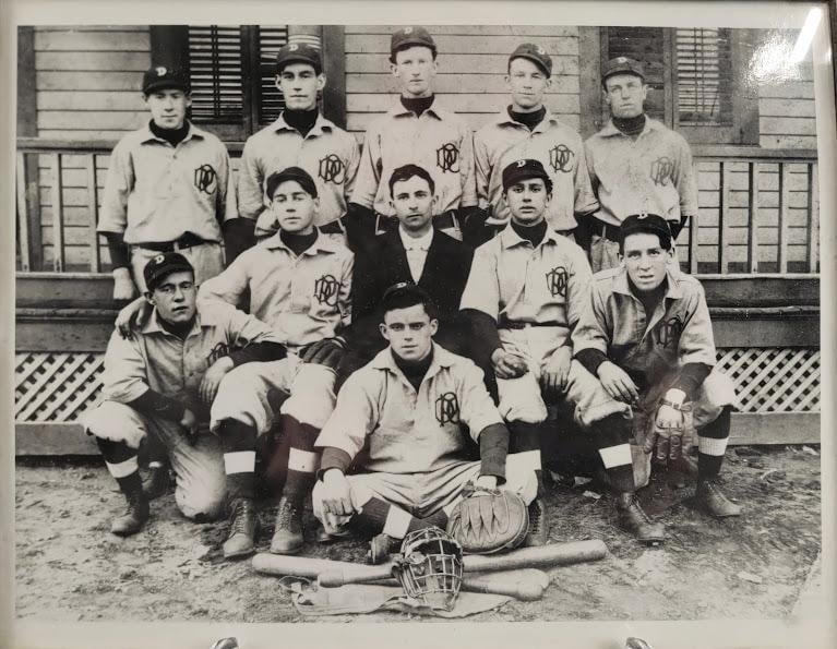 Black-and-white photo of a vintage baseball team posing outdoors in uniform, with gloves and bats on the ground. Eleven men are arranged in three rows in front of a wooden building.