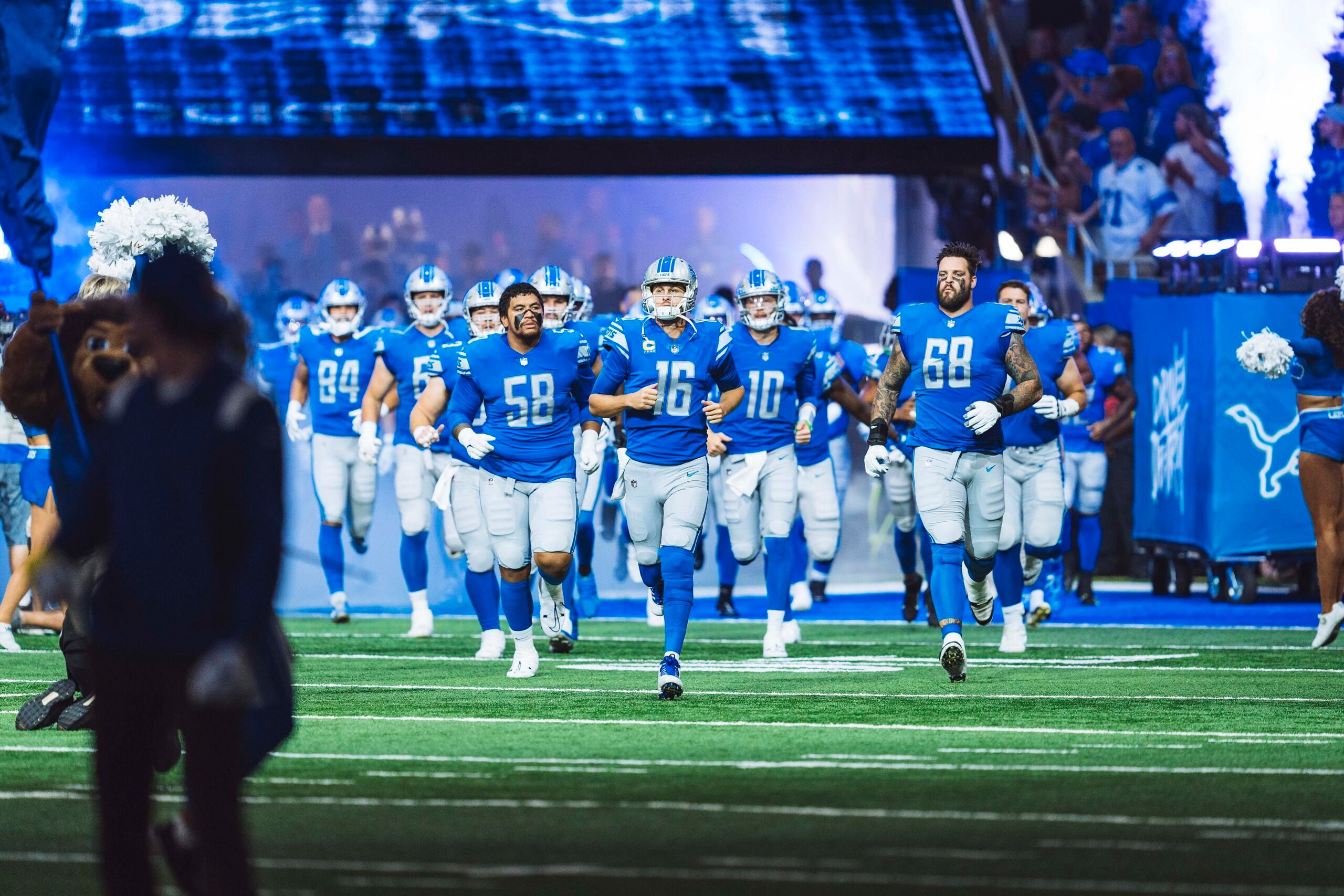 A football team in blue and silver uniforms runs onto a brightly lit field, with cheering fans and cheerleaders welcoming them from the sidelines.