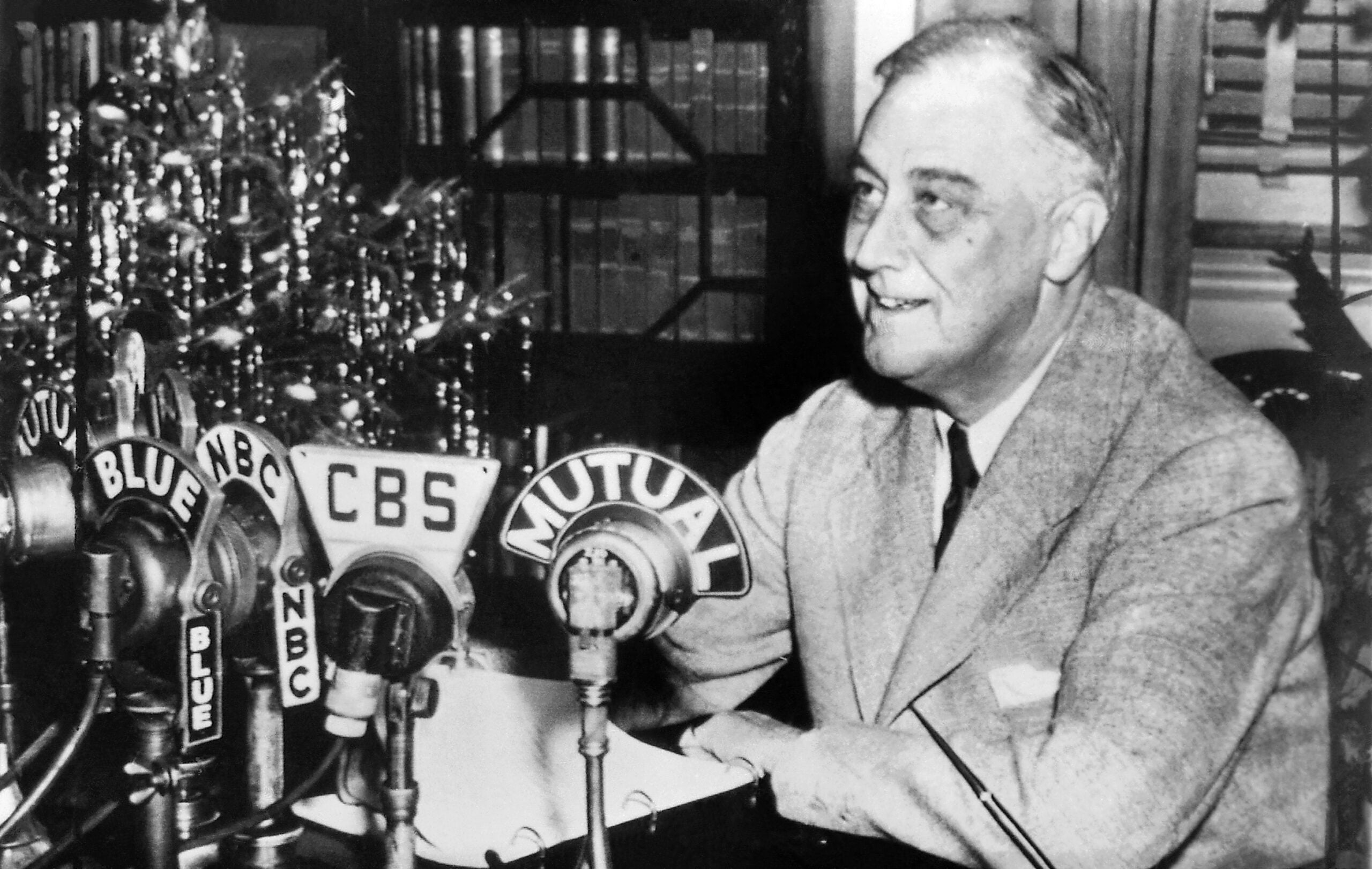 A man in a suit sits at a desk with several vintage microphones labeled CBS, NBC, BLUE, and MUTUAL, smiling while preparing to speak, with bookshelves and a lit tree in the background.