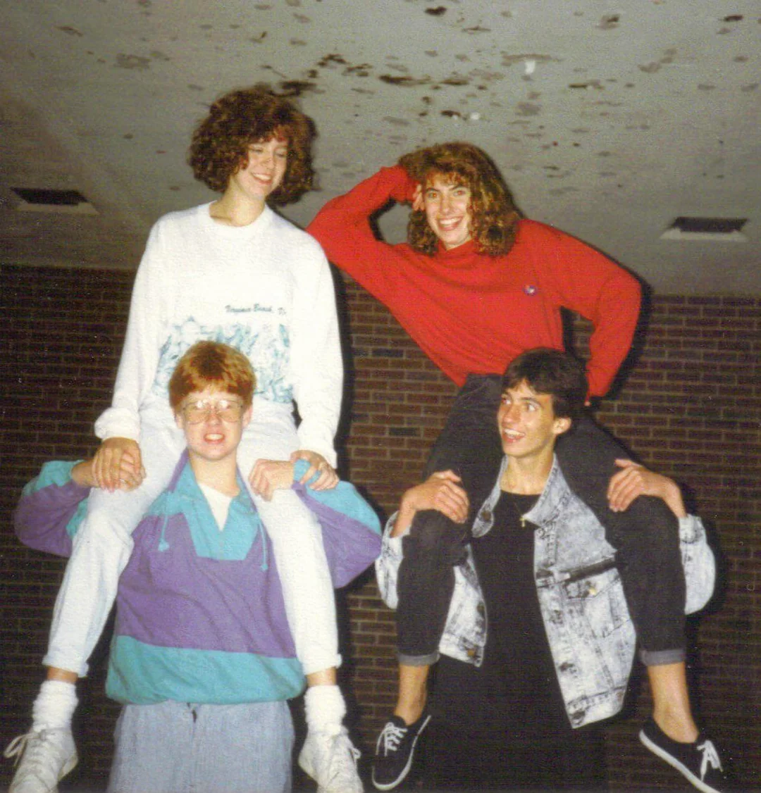 Two smiling women sit on the shoulders of two men. All appear to be teenagers or young adults, posing indoors against a brick wall. The group looks happy and playful.
