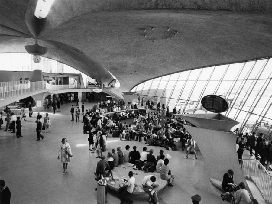 Black-and-white photo of a crowded, futuristic airport terminal with large windows, curved architecture, and people sitting or walking. A central information board and check-in counters are visible.