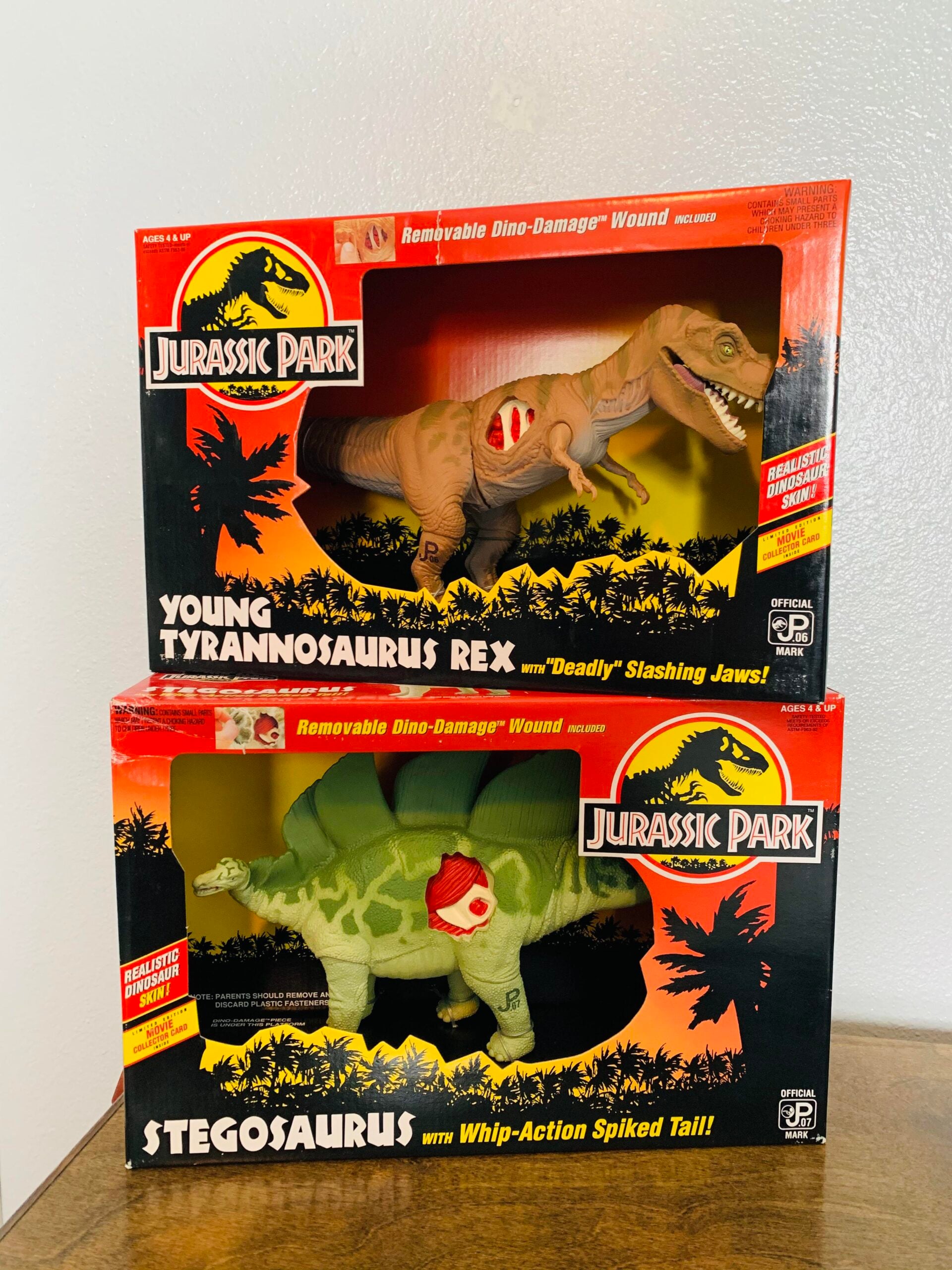 Two Jurassic Park dinosaur toys, Tyrannosaurus Rex and Stegosaurus, in their original red and black boxes with “removable dino-damage” wounds visible on each dinosaur.
