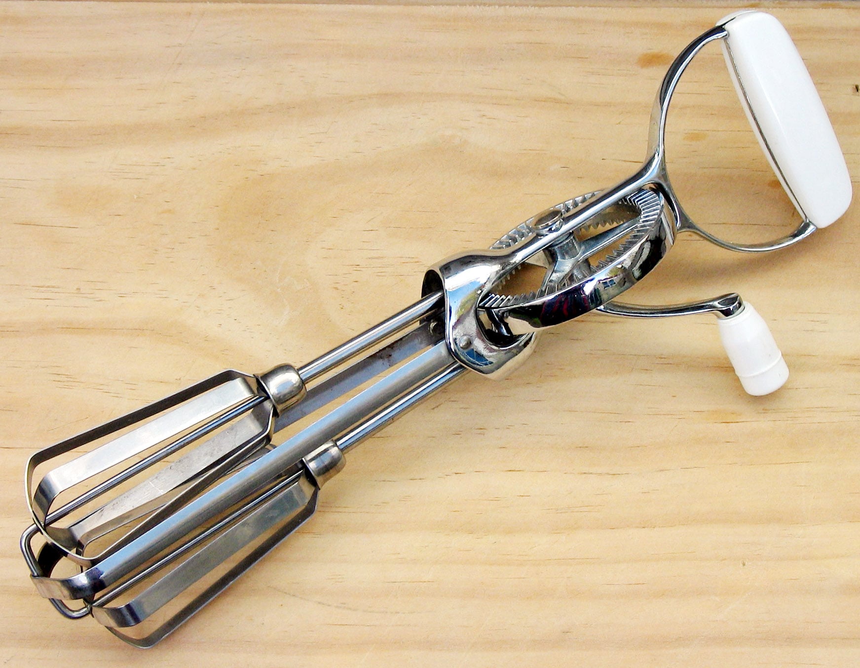 A metal hand-crank egg beater with two beaters and a white handle rests on a light wooden surface.