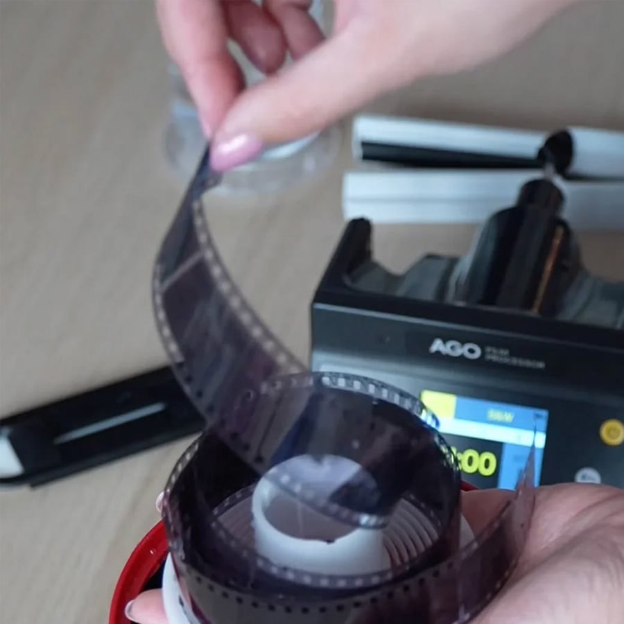 A person loads photographic film onto a developing reel, with a film processing machine and other equipment visible in the background.