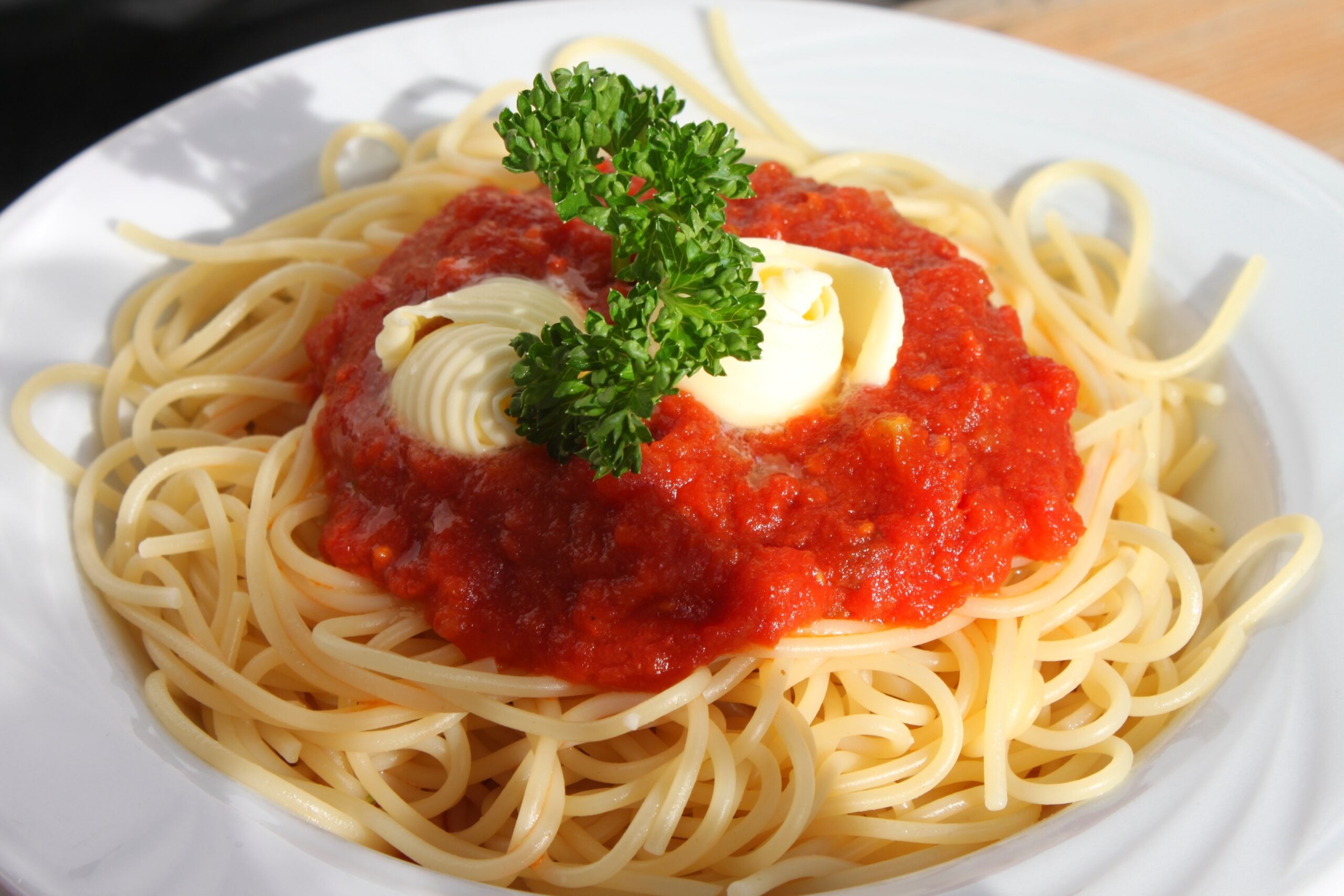 A plate of spaghetti topped with tomato sauce, two curls of butter, and a sprig of parsley, served on a white dish.
