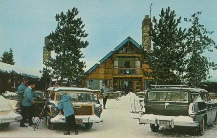 People stand by vintage cars with skis in front of a snowy lodge with a steep roof and stone chimneys, surrounded by pine trees on a winter day.