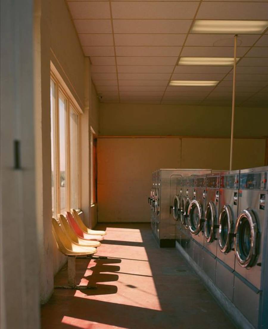 Empty laundromat interior with rows of silver washing machines, yellow plastic chairs beside sunlit windows, and a tile ceiling. Warm sunlight casts long shadows across the floor.