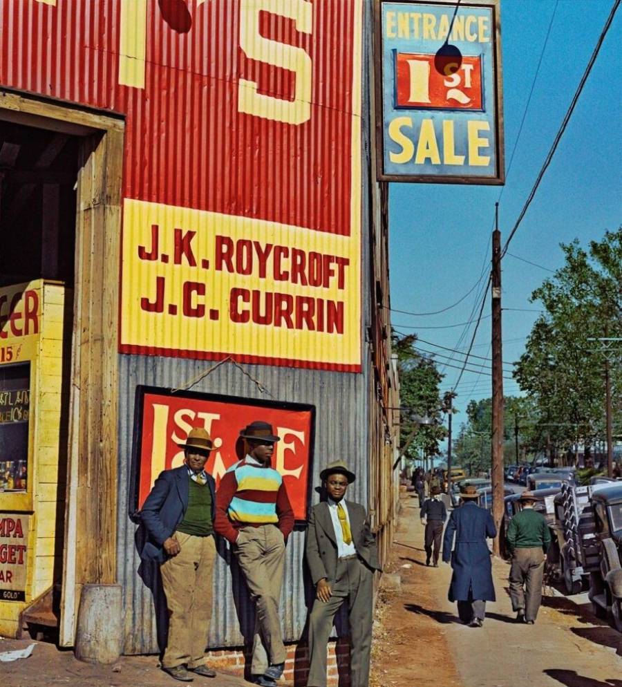 Three men in hats and suits stand by a metal building with large signs reading “J.K. Roycroft” and “J.C. Currin.” People walk along the sidewalk and cars are parked on the street under a clear sky.
