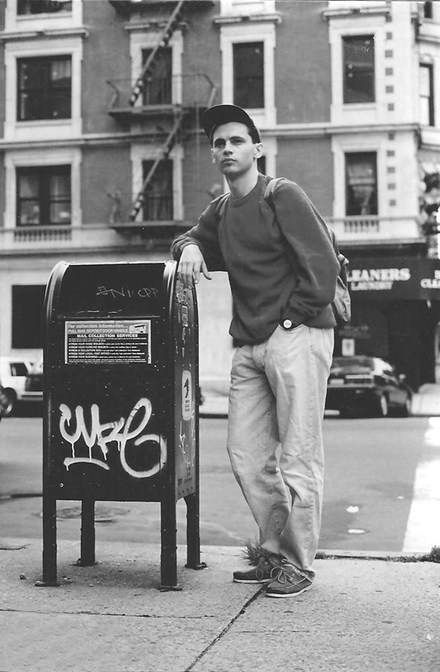 A person in casual clothes leans on a graffiti-covered mailbox on a city sidewalk, with an apartment building and parked cars visible in the background.