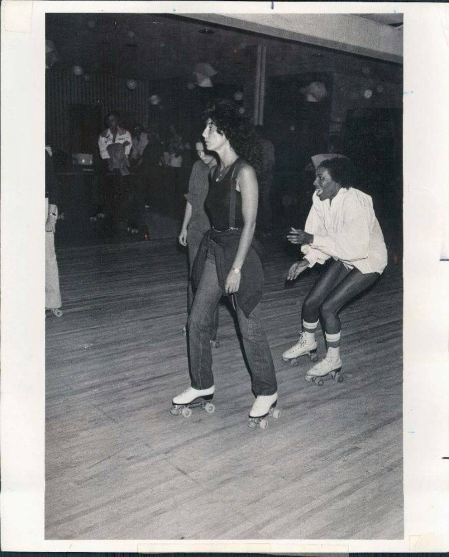Two people roller skating indoors on a wooden rink. One person wears jeans and a vest, the other wears a white shirt and shorts. Other skaters and people are visible in the background. The photo is in black and white.