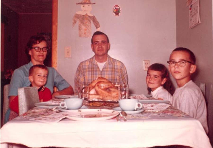 A family of five sits at a dining table with a roast turkey, smiling for a photo. The room is decorated with autumn-themed crafts on the wall.
