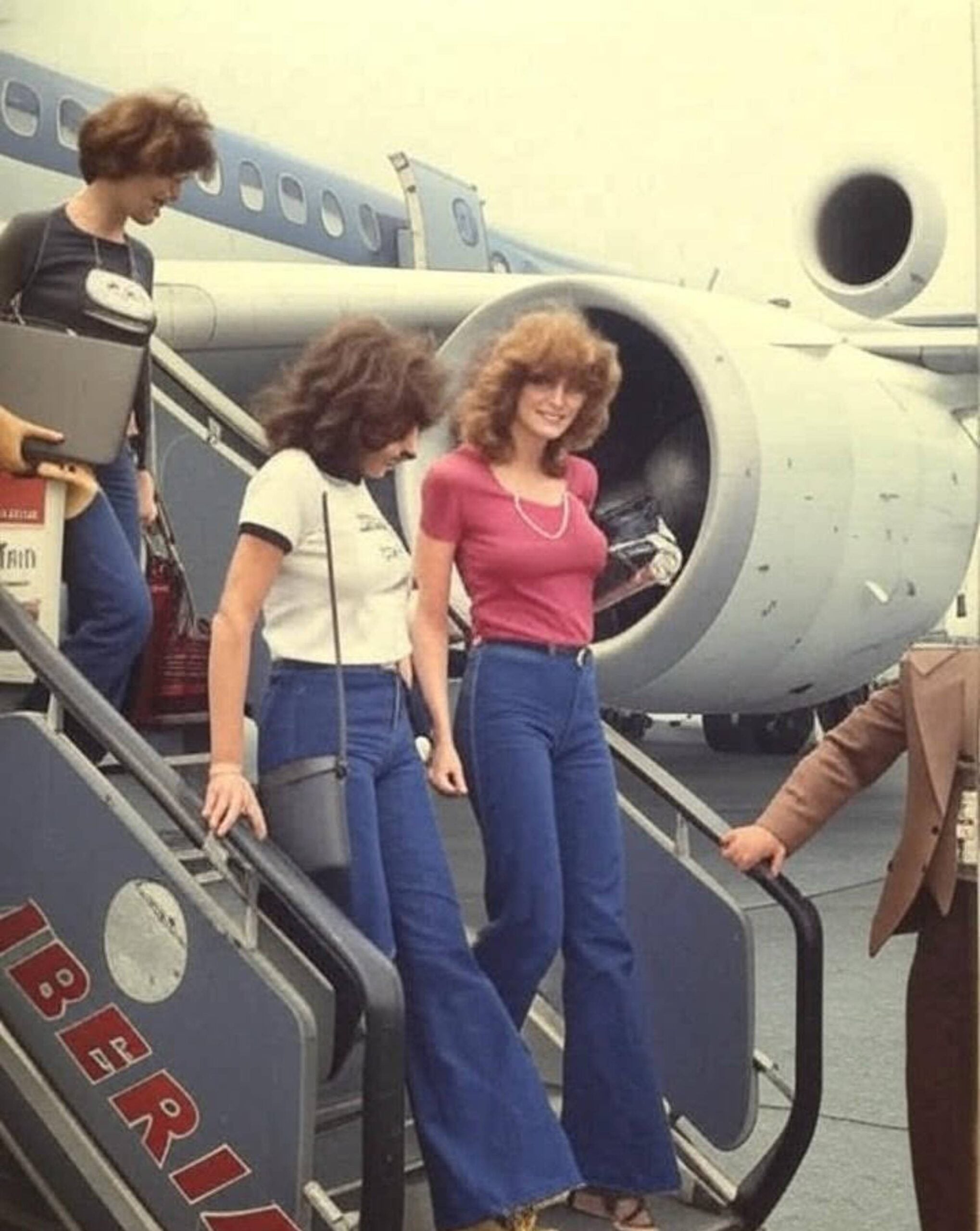 Three women in 1970s outfits descend plane stairs with “IBERIA” written on the side, smiling and talking, with a jet engine visible in the background.