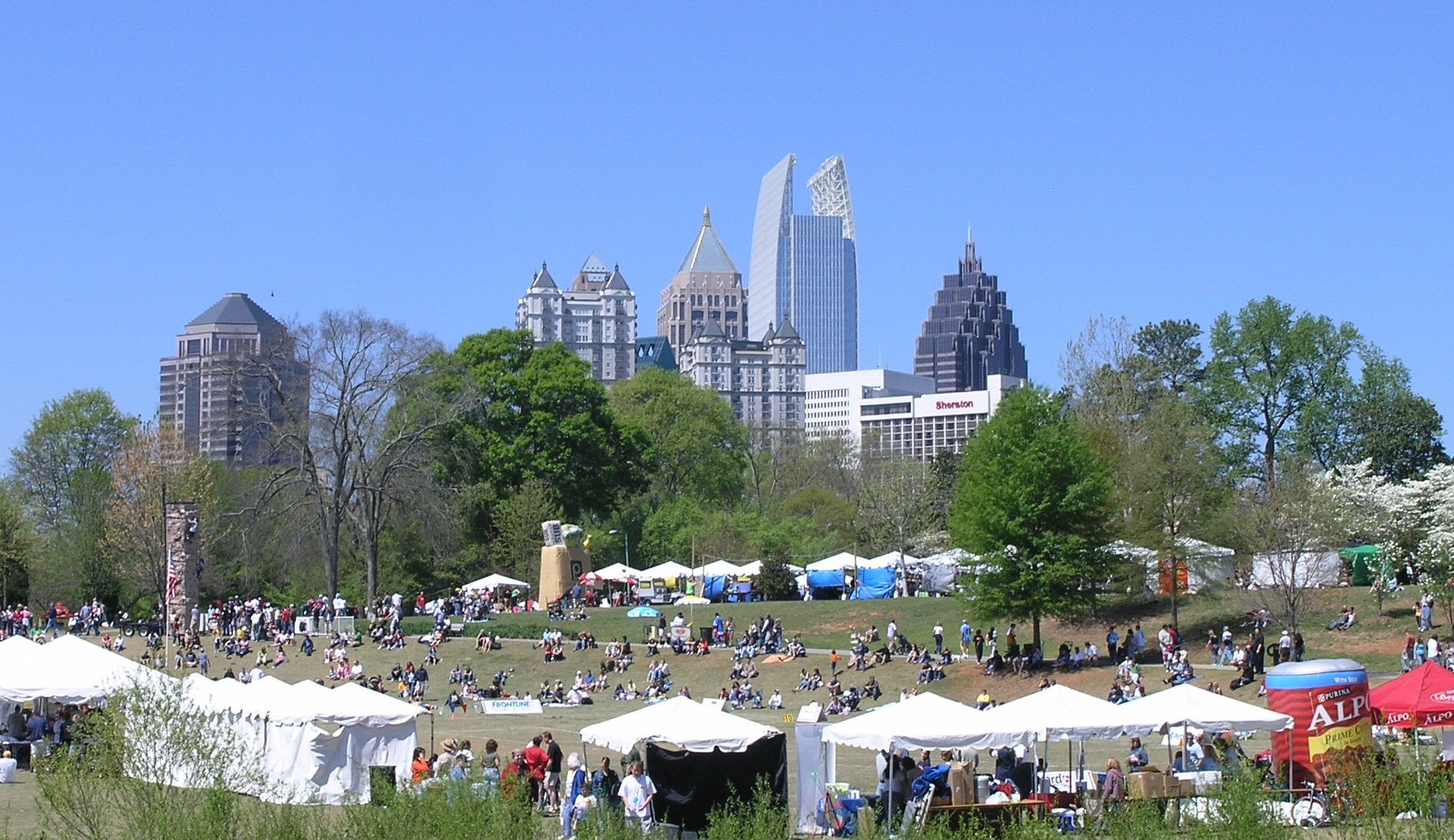 A sunny outdoor festival with white tents, many people on grass, and a skyline of modern and historic buildings in the background, surrounded by green trees under a clear blue sky.