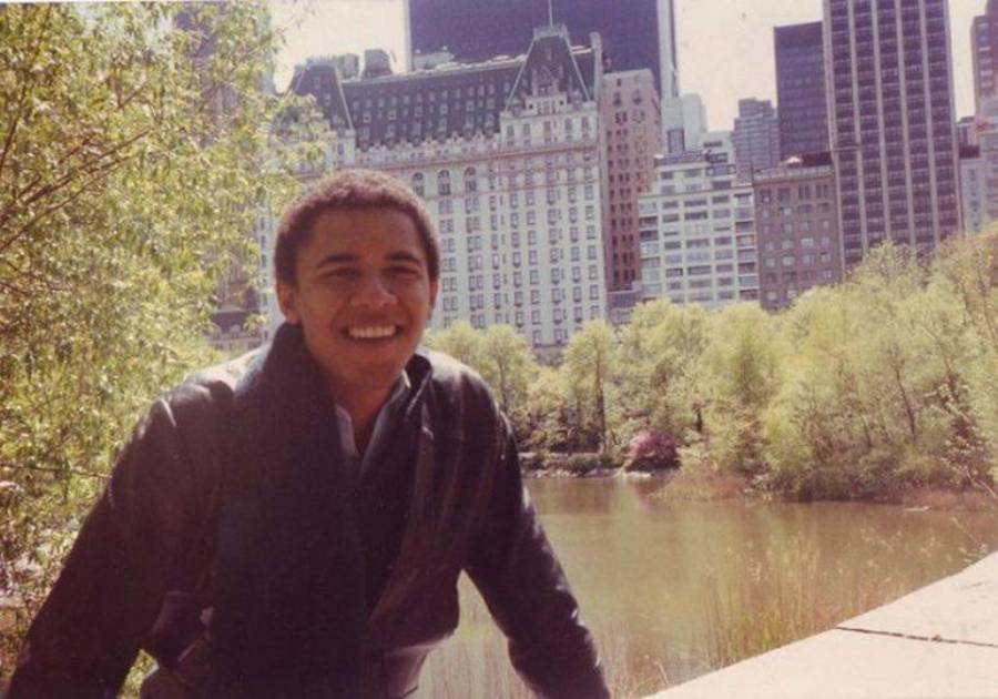 A young man smiles while leaning on a ledge in front of a pond, with tall city buildings and trees in the background on a sunny day.