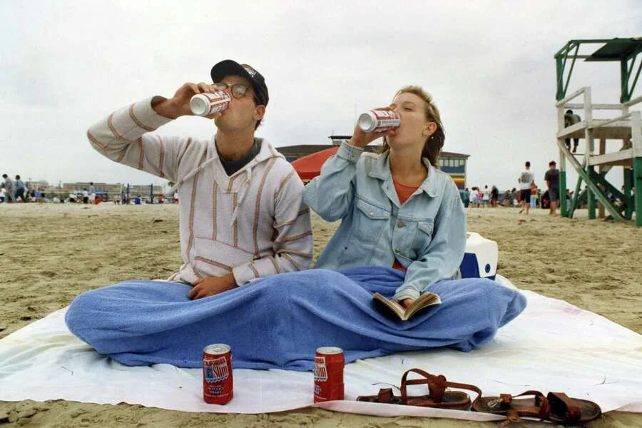 A man and woman sit on a beach under a blue blanket, drinking soda from cans. The woman holds a book. There are sandals, two more soda cans, and people in the background. The sky is cloudy.