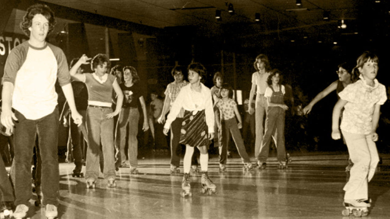 A group of children and teenagers roller skating indoors on a rink, wearing casual 1970s-style clothing. The scene is lively and the image has a sepia tone.