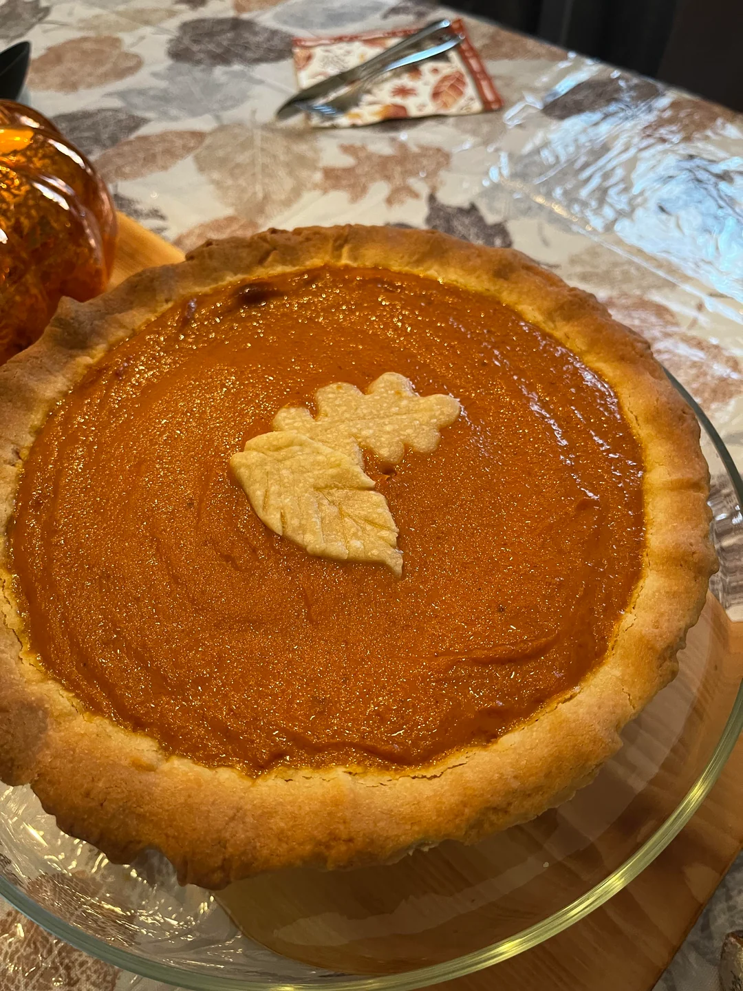 Pumpkin pie with a golden-brown crust, topped with a decorative pastry leaf, sits on a glass pie dish on a table with a fall-themed tablecloth.