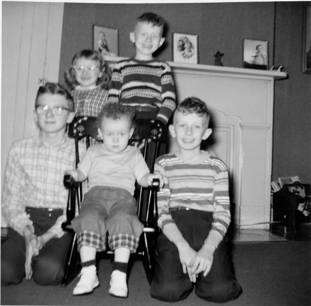 Five children pose for a photo indoors, with one child seated in a small chair. Three framed photos and decorative items are visible on the mantel behind them. The image is in black and white.