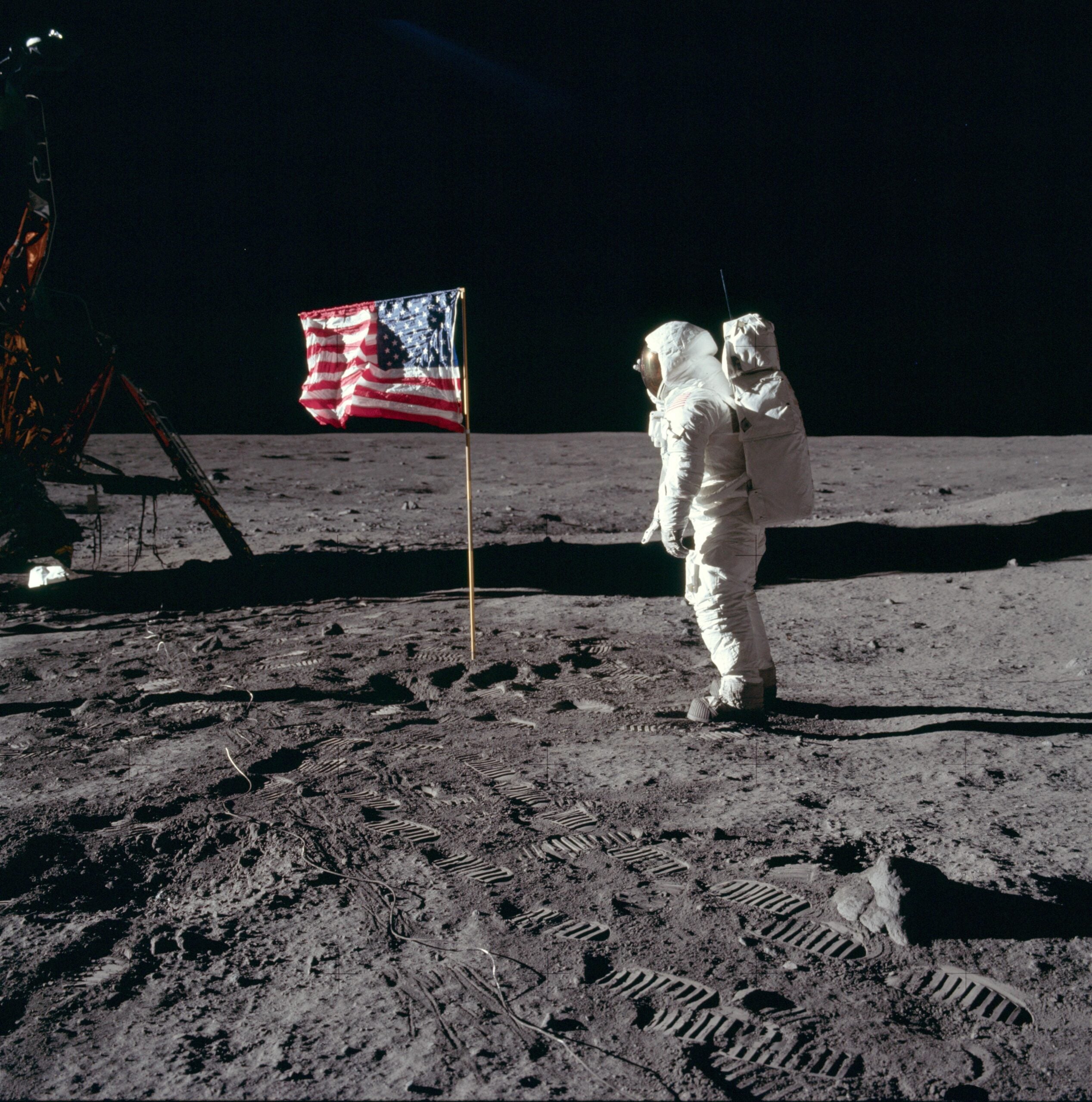 An astronaut in a spacesuit stands on the Moon near an American flag, with the lunar module visible on the left and footprints in the gray, dusty surface. The sky above is completely black.