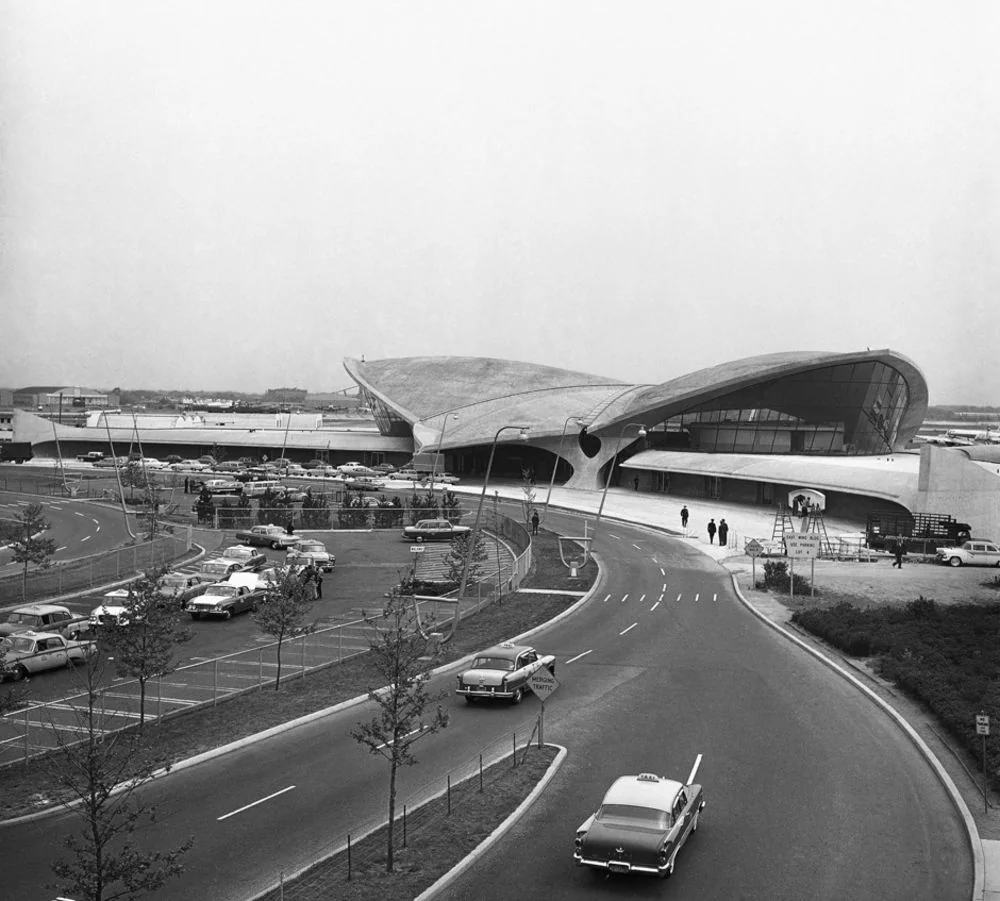 Black and white photo of a futuristic airport terminal with sweeping curves, vintage cars on a road and parked, and people walking near the entrance. The scene appears to be from the mid-20th century.