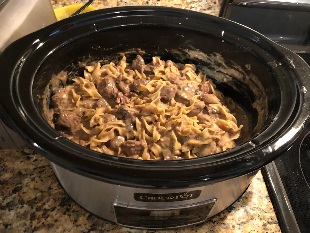 A crockpot filled with cooked egg noodles, beef, and creamy sauce sits on a granite kitchen counter.