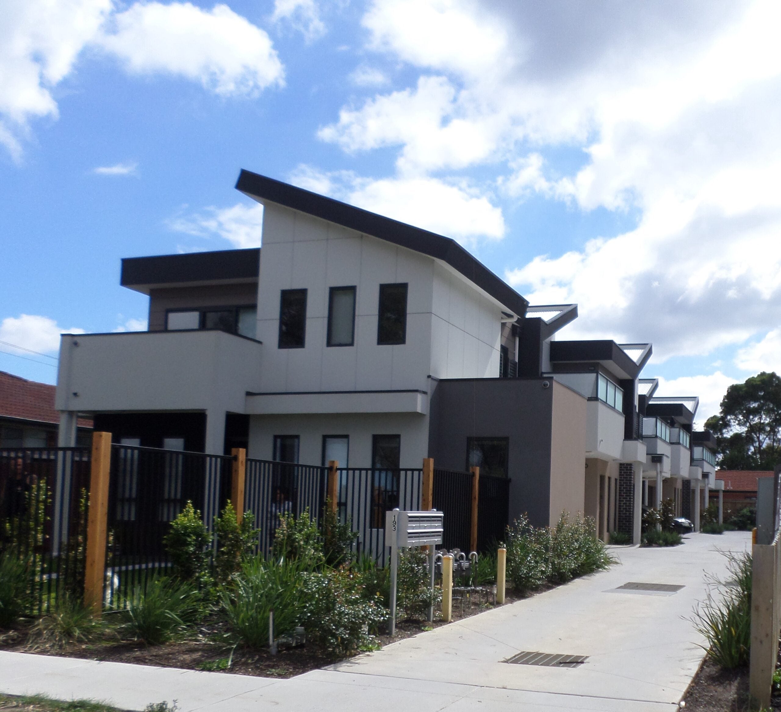 Modern two-story townhouses with large windows, angular roofs, a black fence, and landscaped greenery along a paved pathway under a partly cloudy sky.