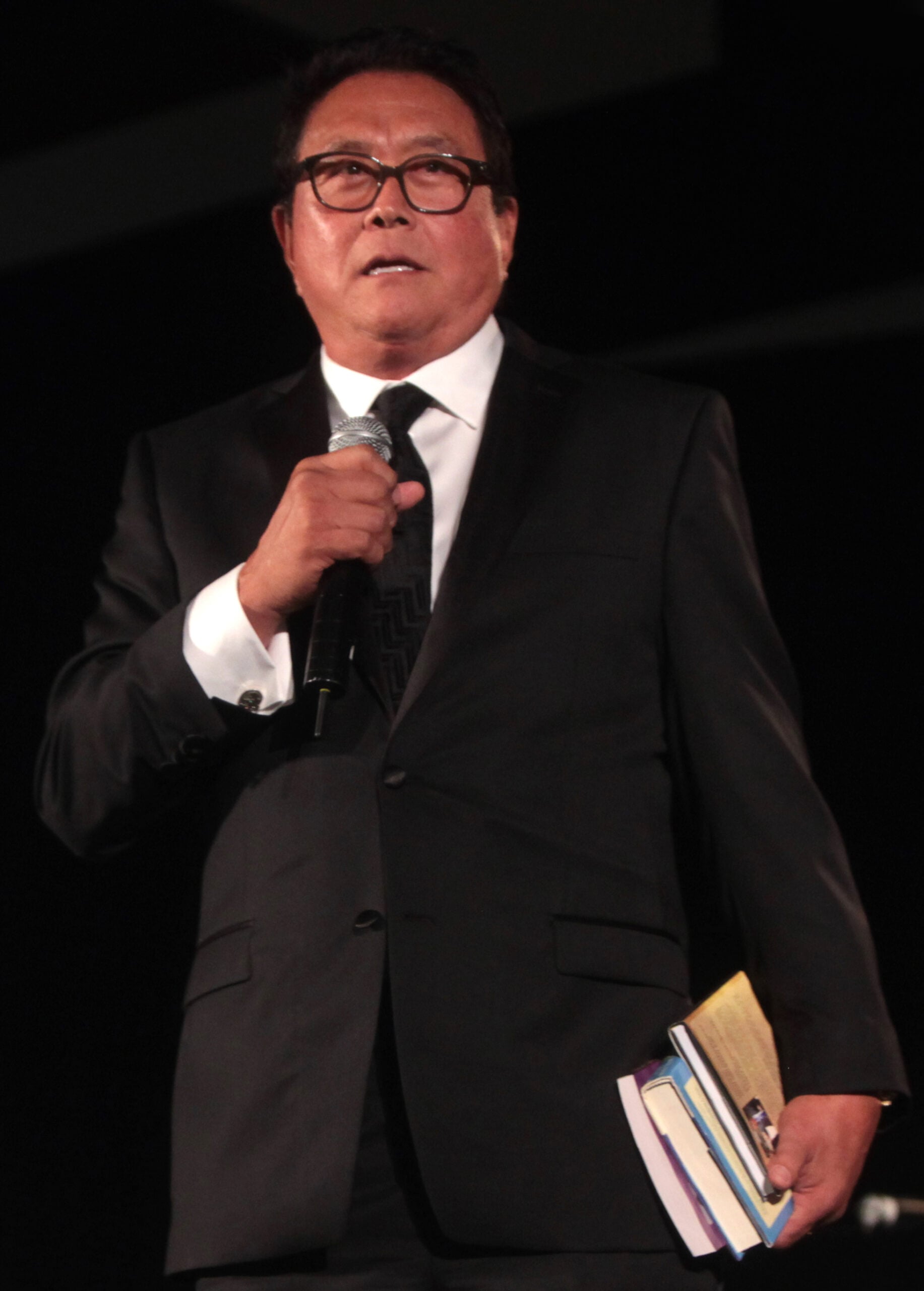 A man in a black suit and glasses holds a microphone and several books while standing on stage against a dark background.