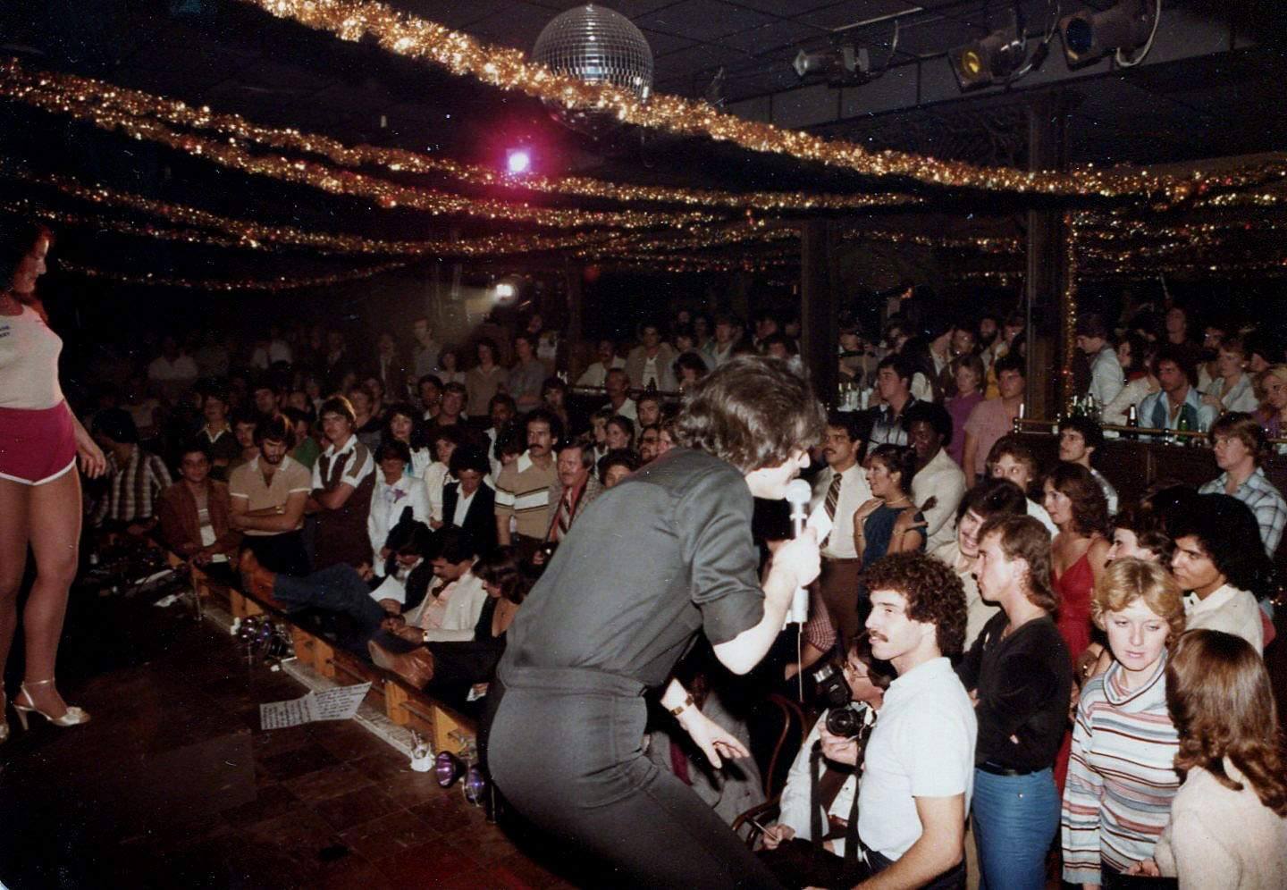 A crowded nightclub with a performer singing on stage, people seated and standing, watching attentively. Gold tinsel decorations hang from the ceiling, and a disco ball is visible above the audience.