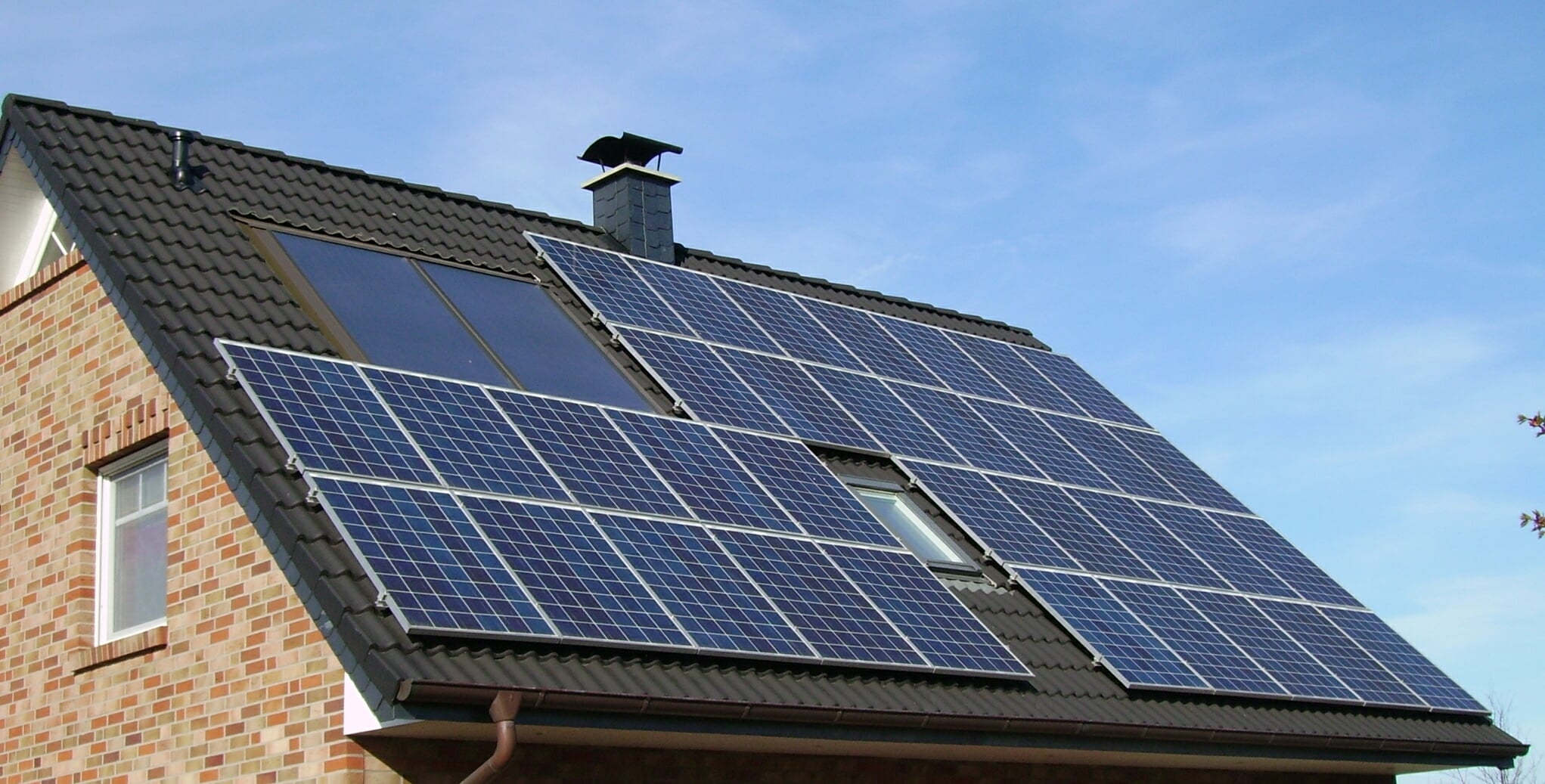 A house with a steep, tiled roof has several solar panels installed, with a blue sky in the background. The building is made of red and tan bricks.