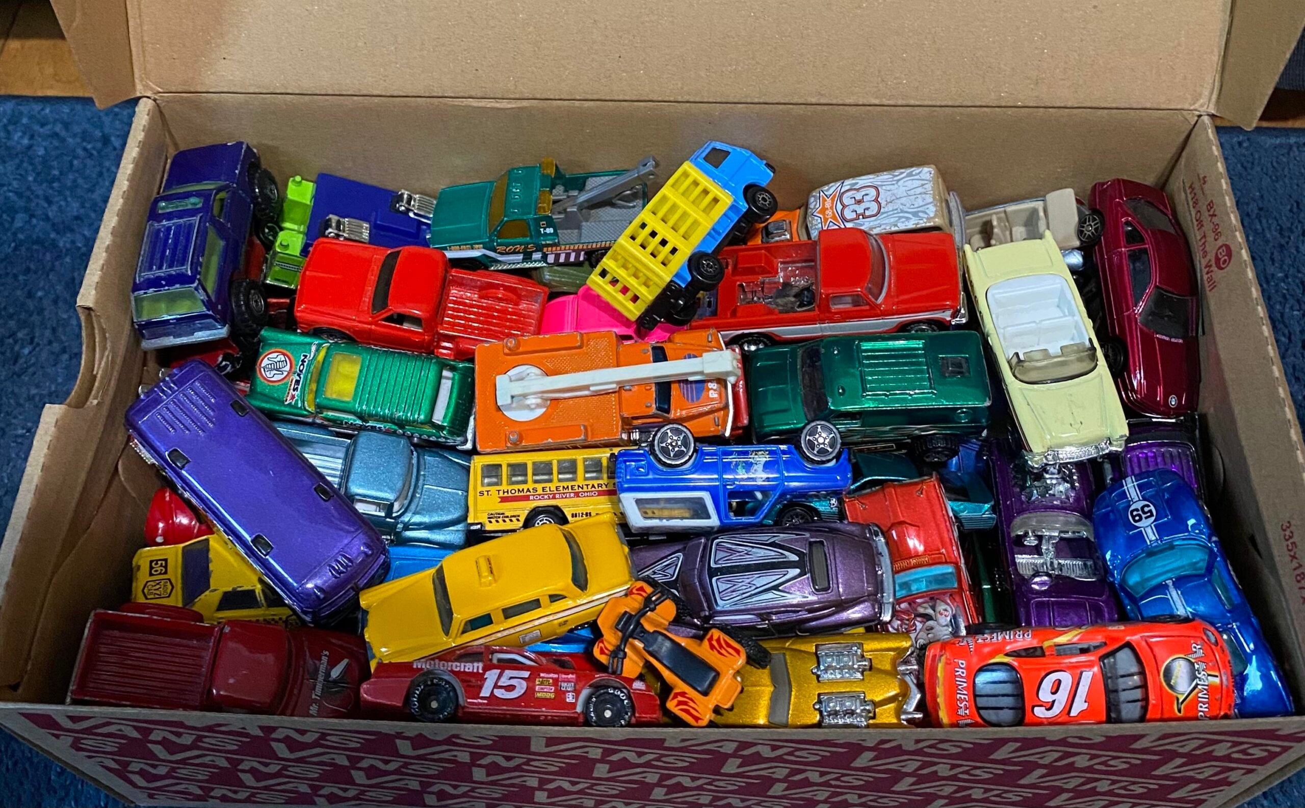A cardboard Vans shoe box filled with assorted colorful toy cars, trucks, and buses, all jumbled together on a blue carpeted floor.