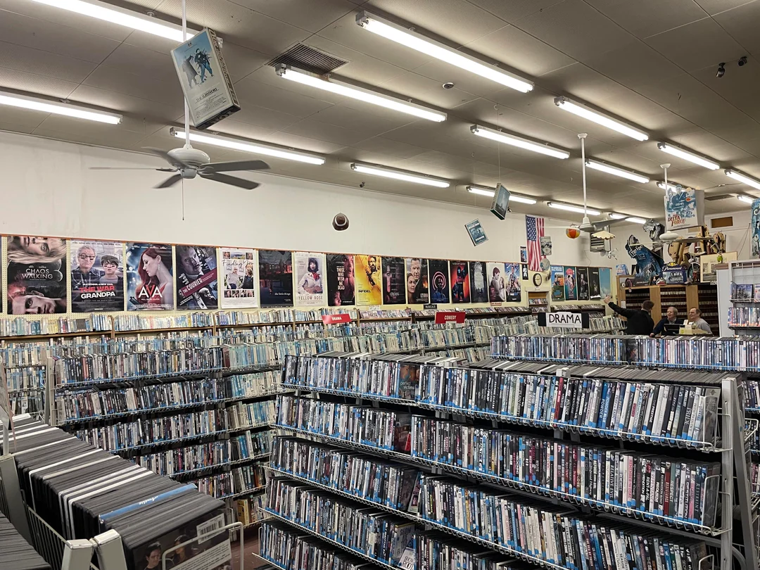 Rows of DVDs fill a store under bright lights. Movie posters line the walls. Hanging decorations and signs, such as “Drama,” are visible above the shelves. A few people browse in the background.