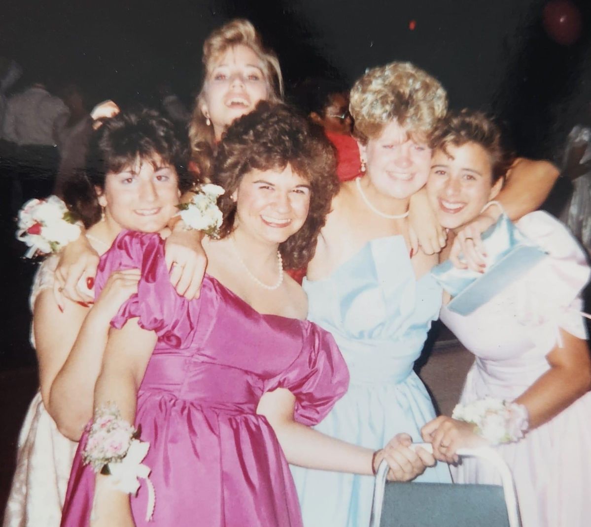 Five young women in colorful 1980s prom dresses smile and pose together, some with arms around each other. They are wearing corsages and appear happy, standing closely in a festive indoor setting.