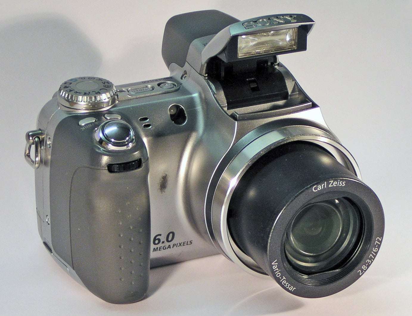 A silver digital camera with a protruding lens and a raised flash. The lens displays “Carl Zeiss” branding and “6.0 mega pixels” is labeled on the camera body.