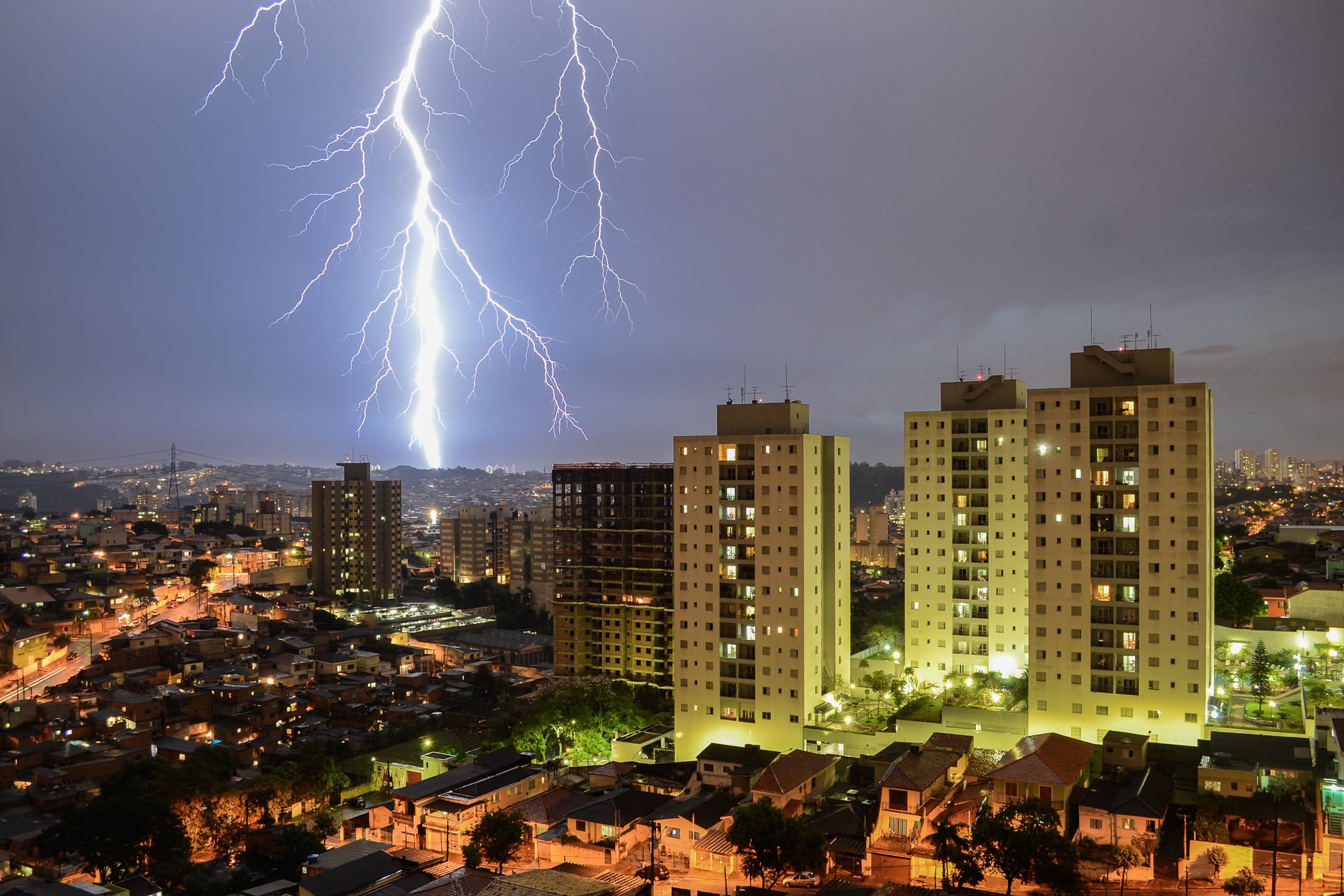 Bright lightning strikes in the night sky above a city with tall apartment buildings and many lights, highlighting a dramatic urban landscape.