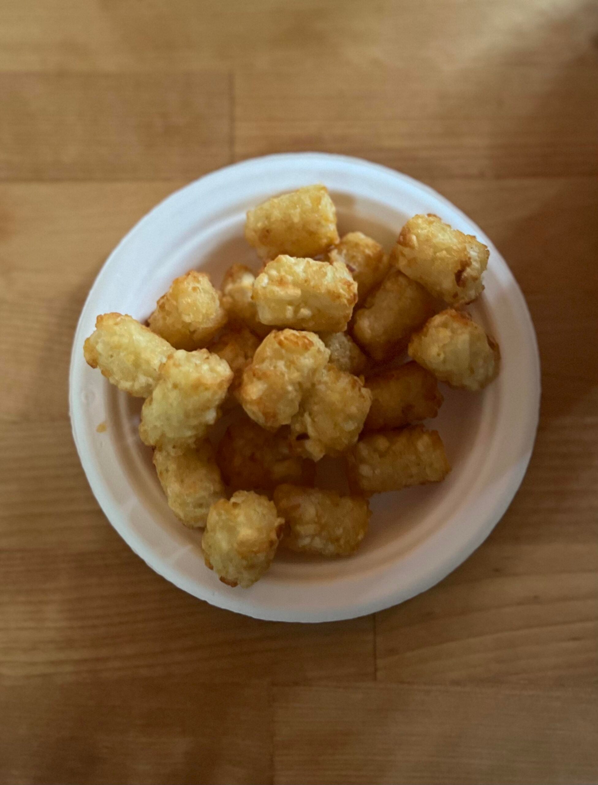A white bowl filled with golden, crispy tater tots sits on a wooden surface.